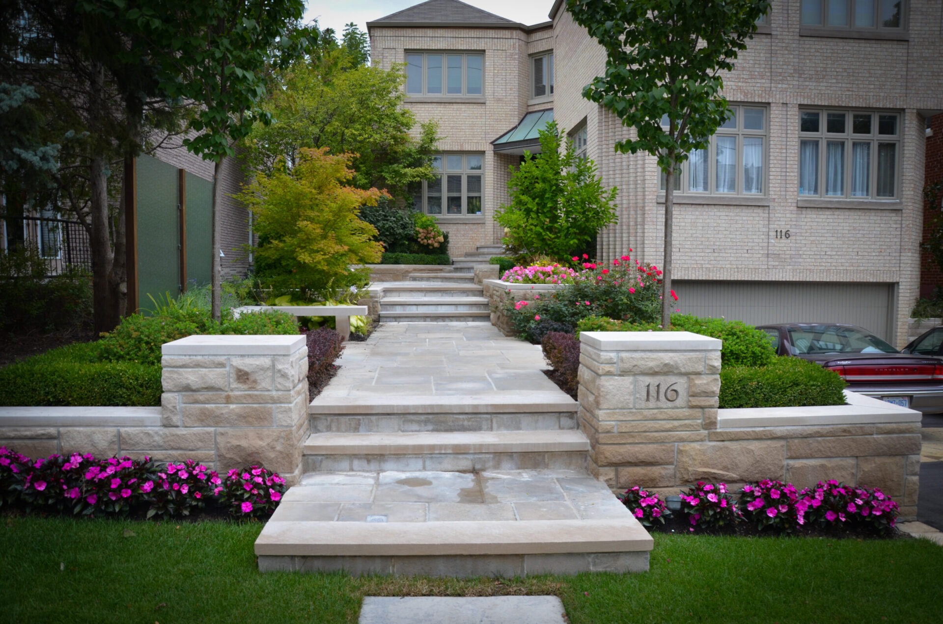 Elegant residential entrance with stone steps, manicured bushes, and vibrant flowers leading to a modern, two-story brick building surrounded by trees.