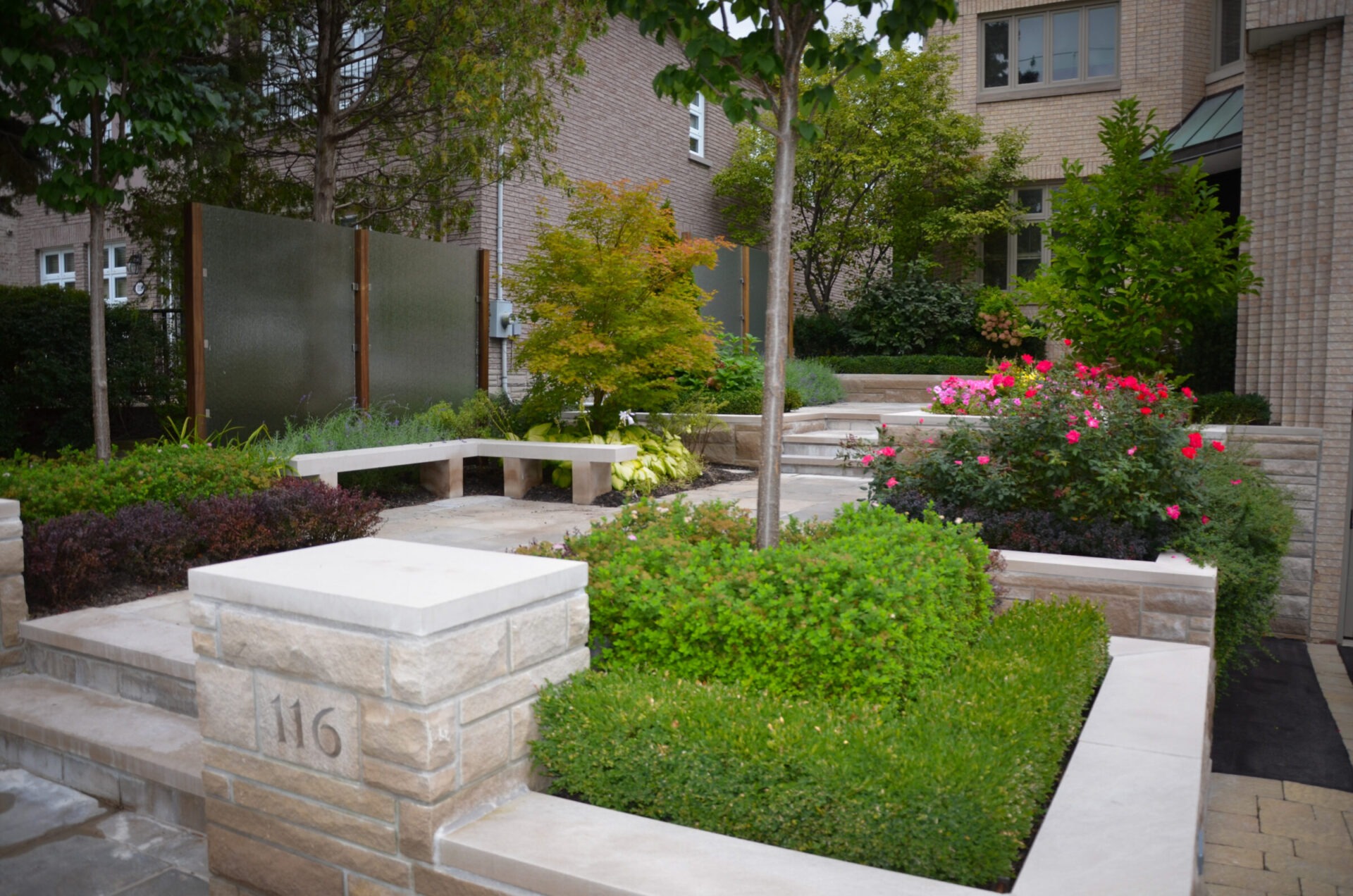 A small urban garden with stone benches, lush greenery, and flowering shrubs, surrounded by beige brick buildings. Address 