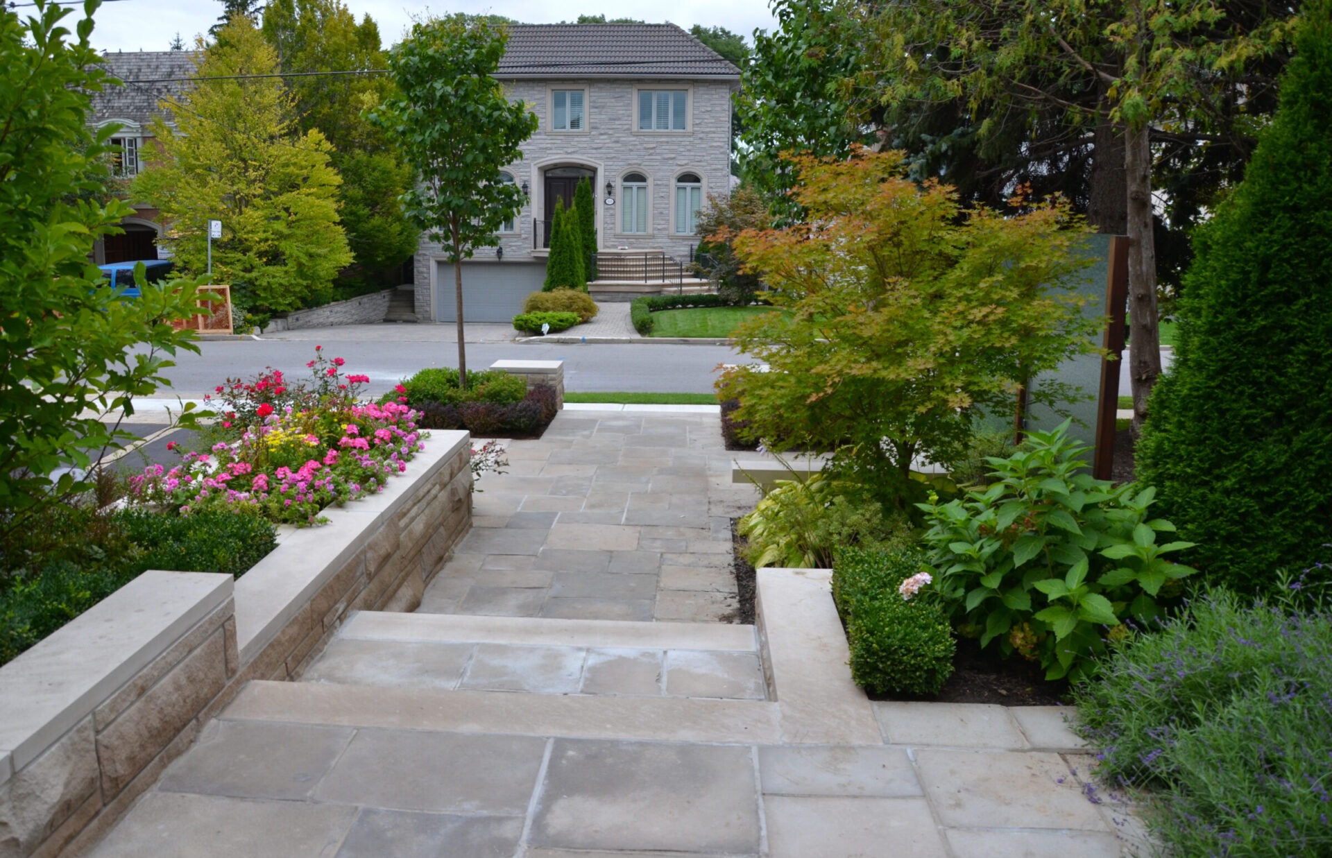 A stone path with landscaped gardens leads to a large house on a residential street, surrounded by trees and lush greenery.