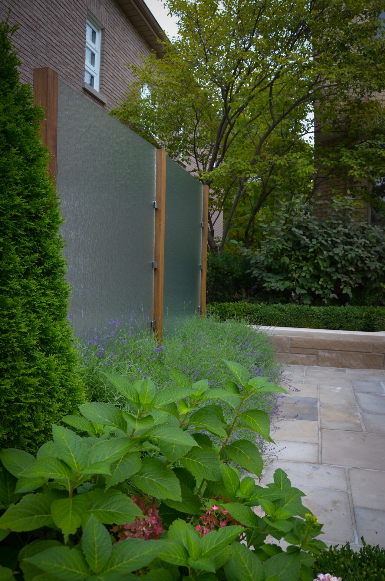 A serene garden features lush green plants, a modern frosted glass privacy screen, and a charming brick building amidst leafy trees.