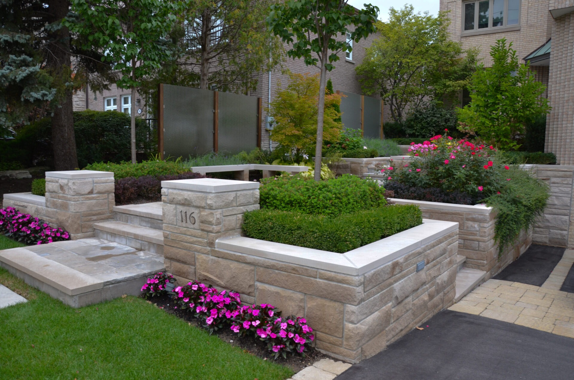 A modern residential garden features neatly arranged stone walls, lush greenery, colorful flowers, and hedges, providing an elegant, structured outdoor space.