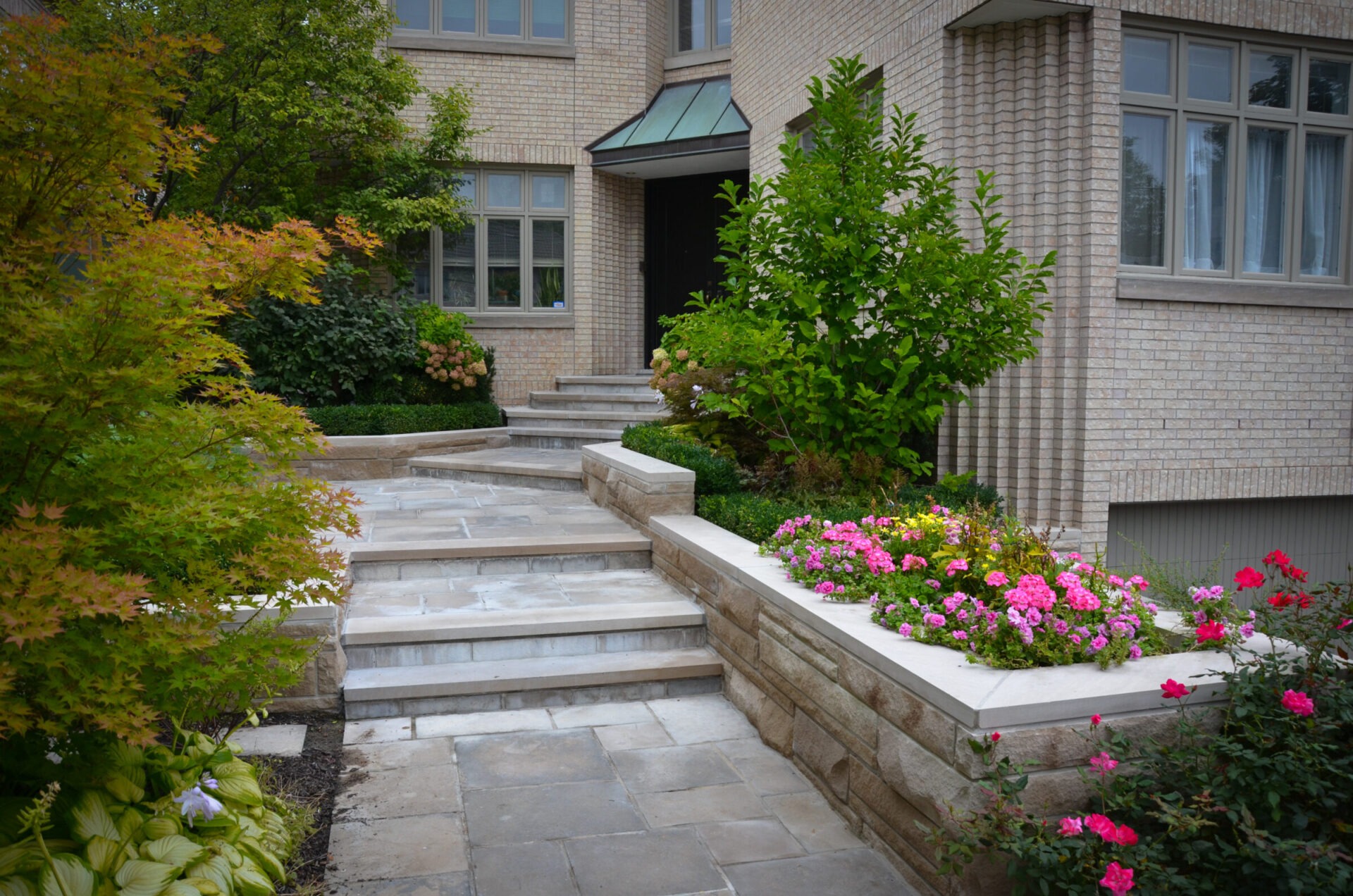 Brick house facade with stone pathway and steps, lush greenery, and vibrant flowerbeds creating a welcoming entrance.