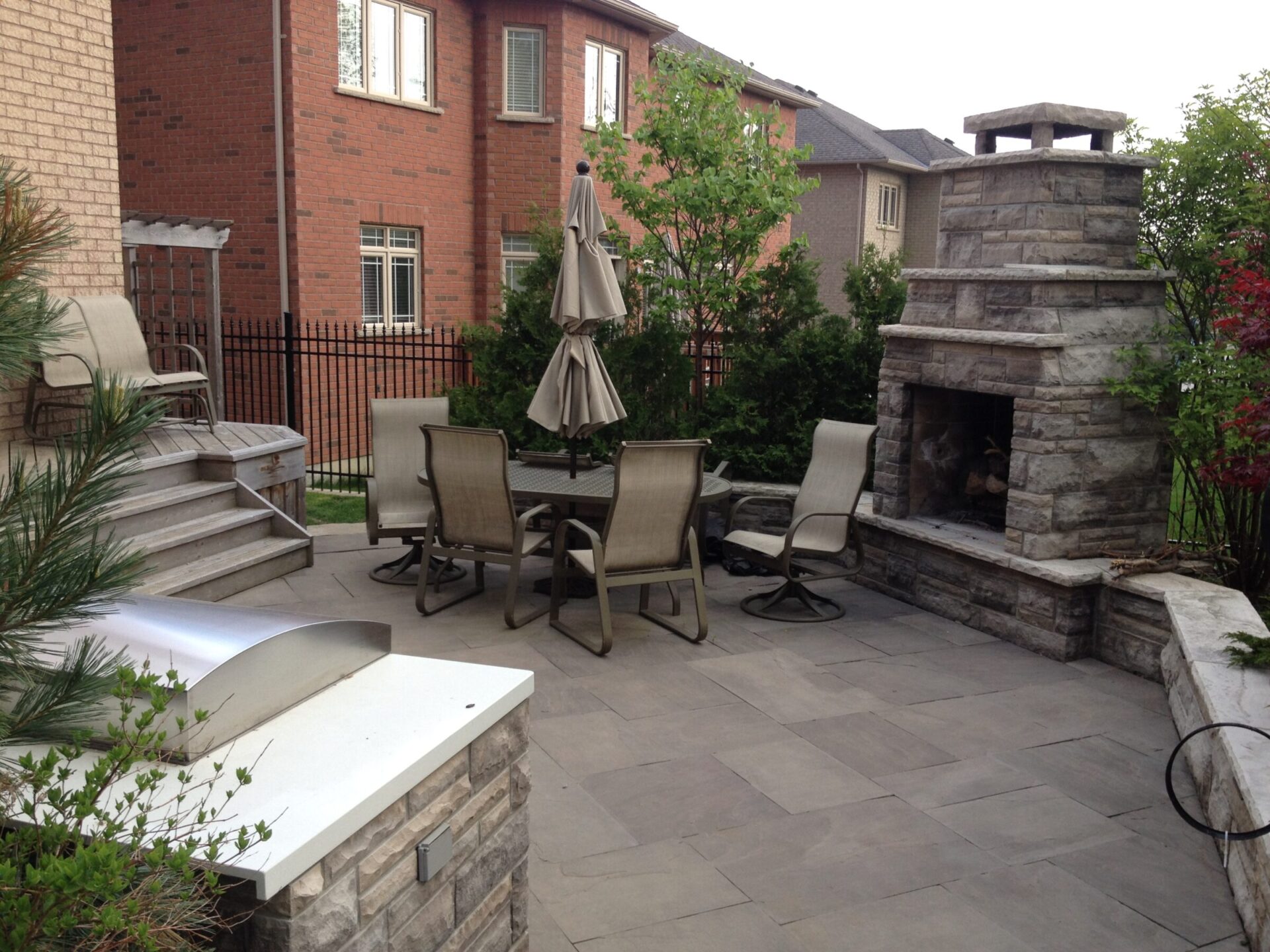 A cozy backyard patio features stone flooring, a round table with chairs, an umbrella, and a stone fireplace surrounded by brick houses and greenery.