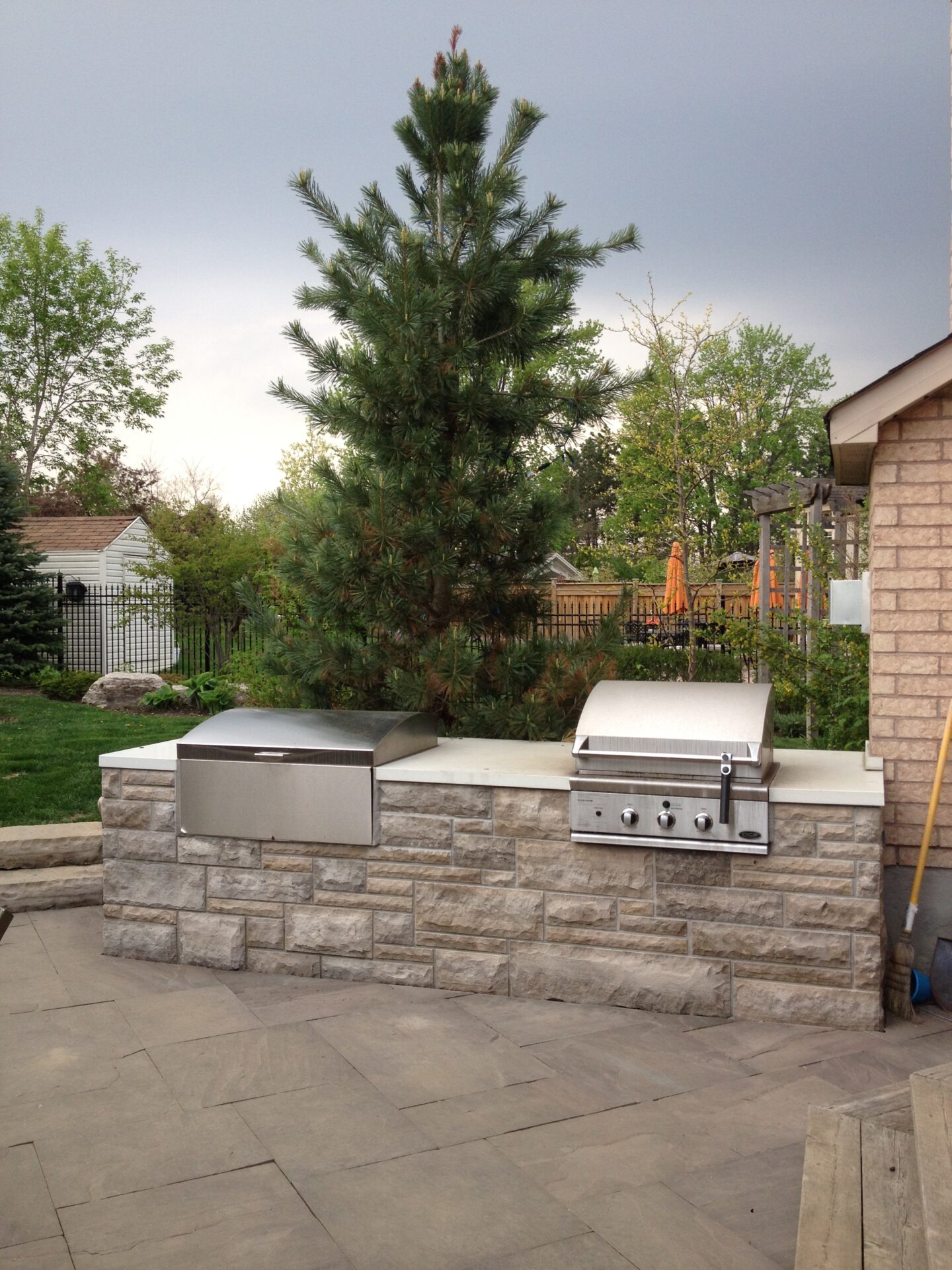 Outdoor patio with a stone grill and counter, set against a backdrop of green trees and a cloudy sky. Cozy garden setting.