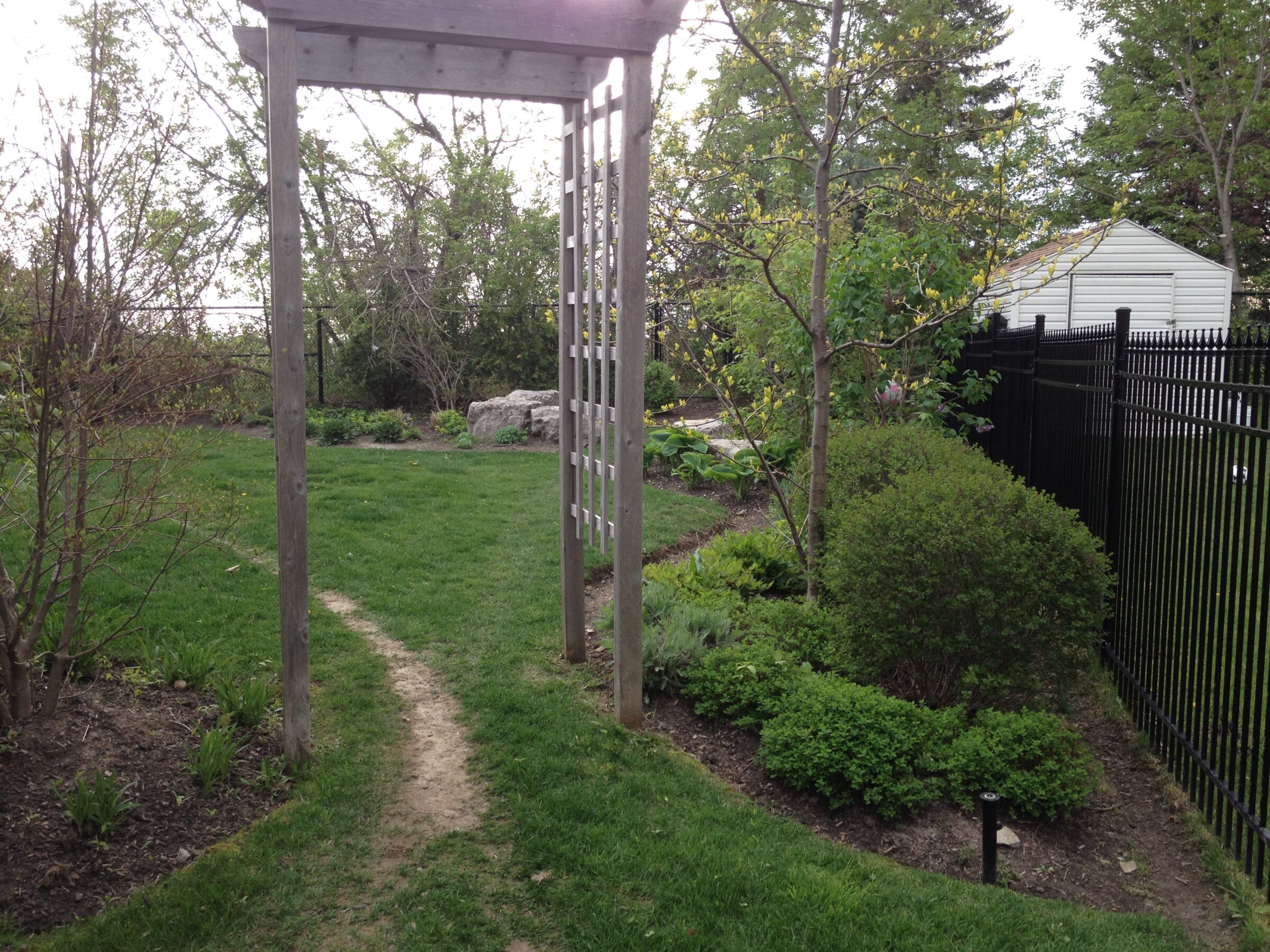 A green garden path leads through a wooden arbor surrounded by lush vegetation and trees. A black fence lines the right side.