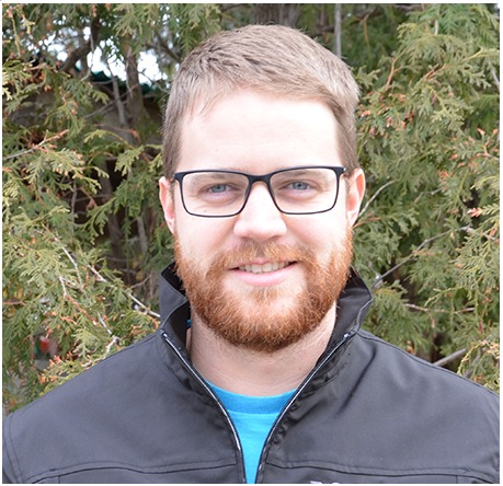 A person with glasses stands in front of green foliage, wearing a black jacket and blue shirt, smiling at the camera.