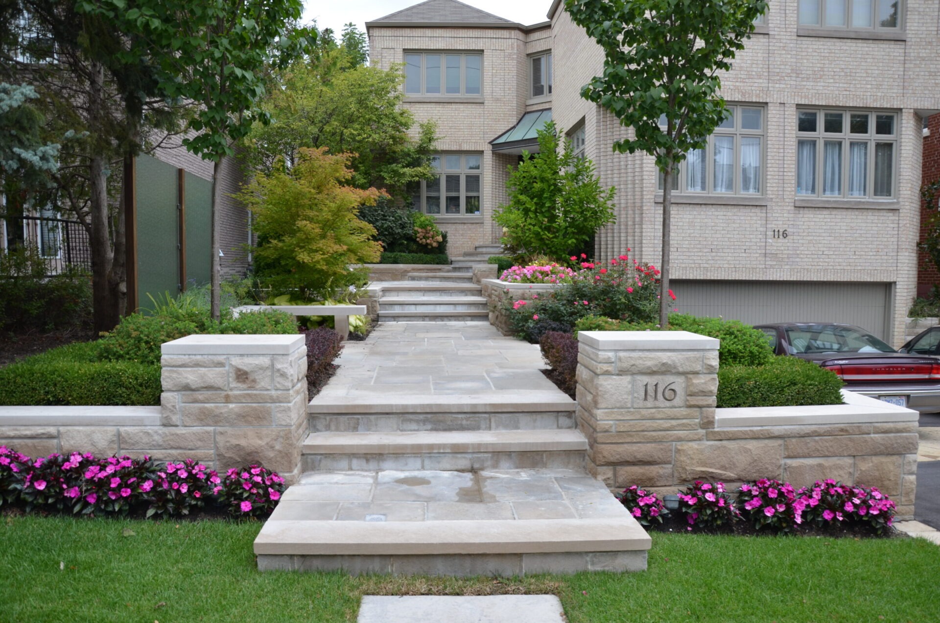 Two-story suburban house with a stone driveway, well-maintained garden, and vibrant flowers. Steps lead to entrance, flanked by lush greenery.