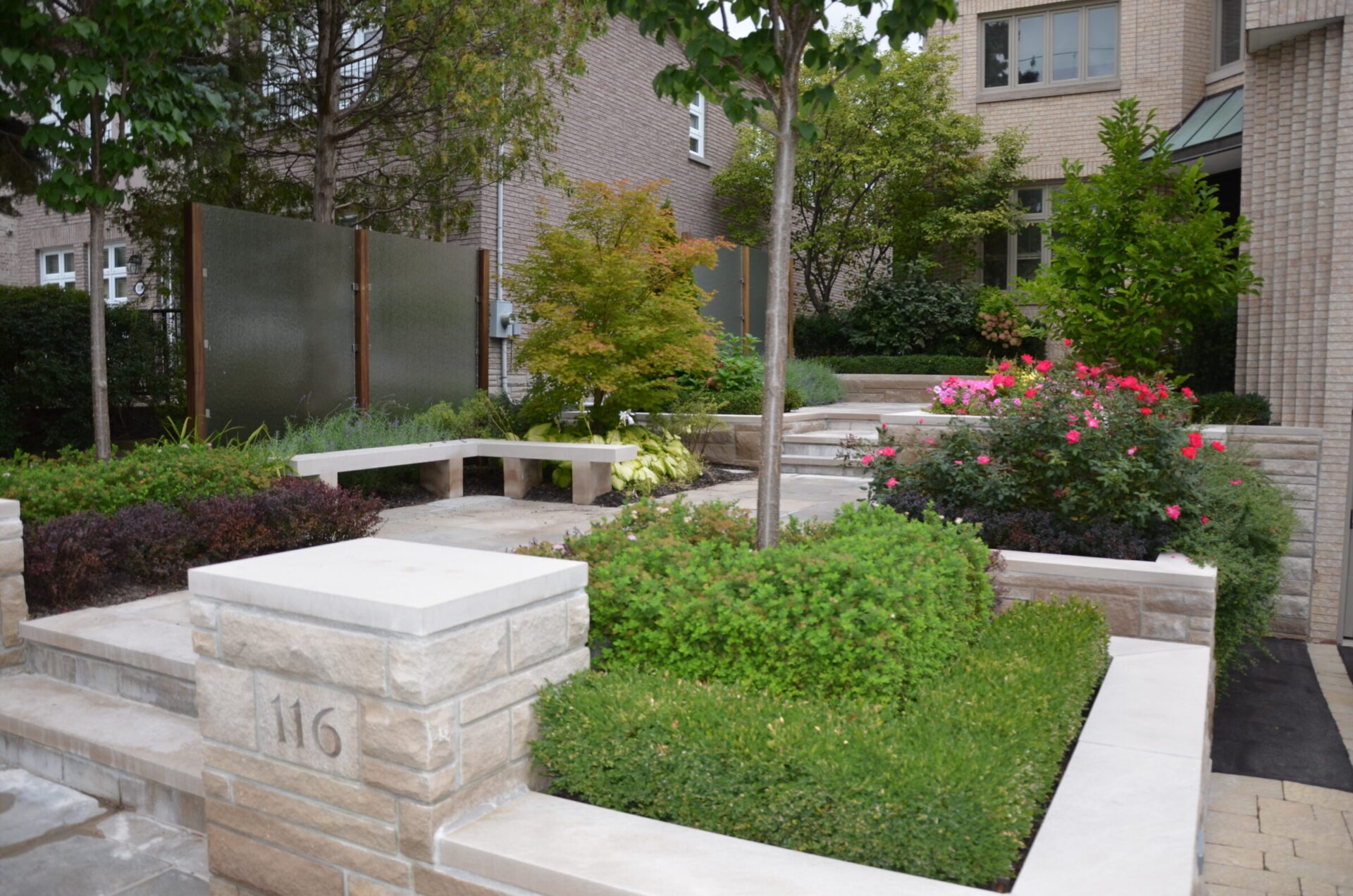 Modern garden with neatly trimmed bushes and vibrant flowers, surrounded by stone walls. Brick buildings in the background provide an urban setting.