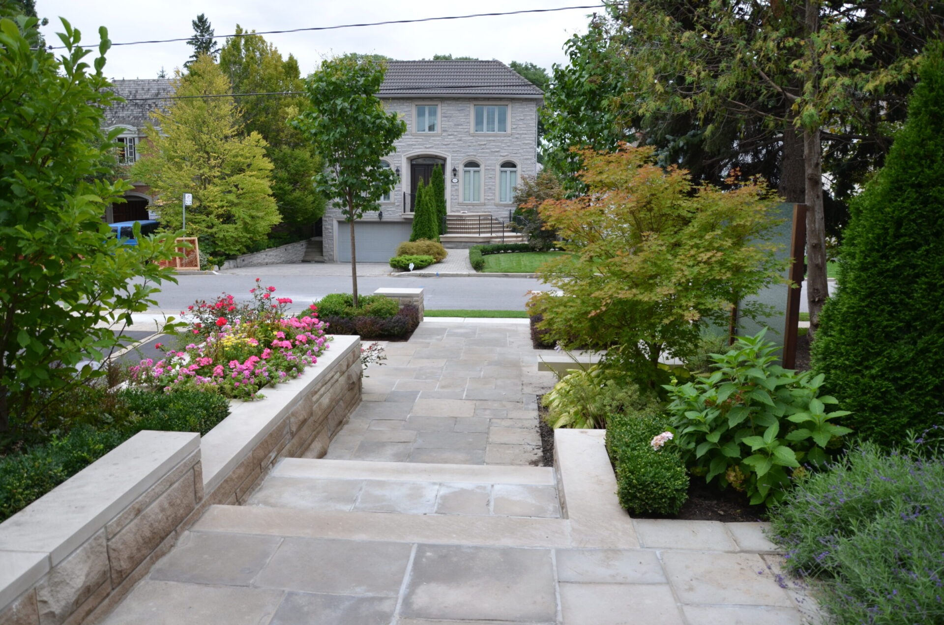 A stone pathway and steps lead to a well-manicured garden with lush greenery and flowers, facing a two-story residential house.