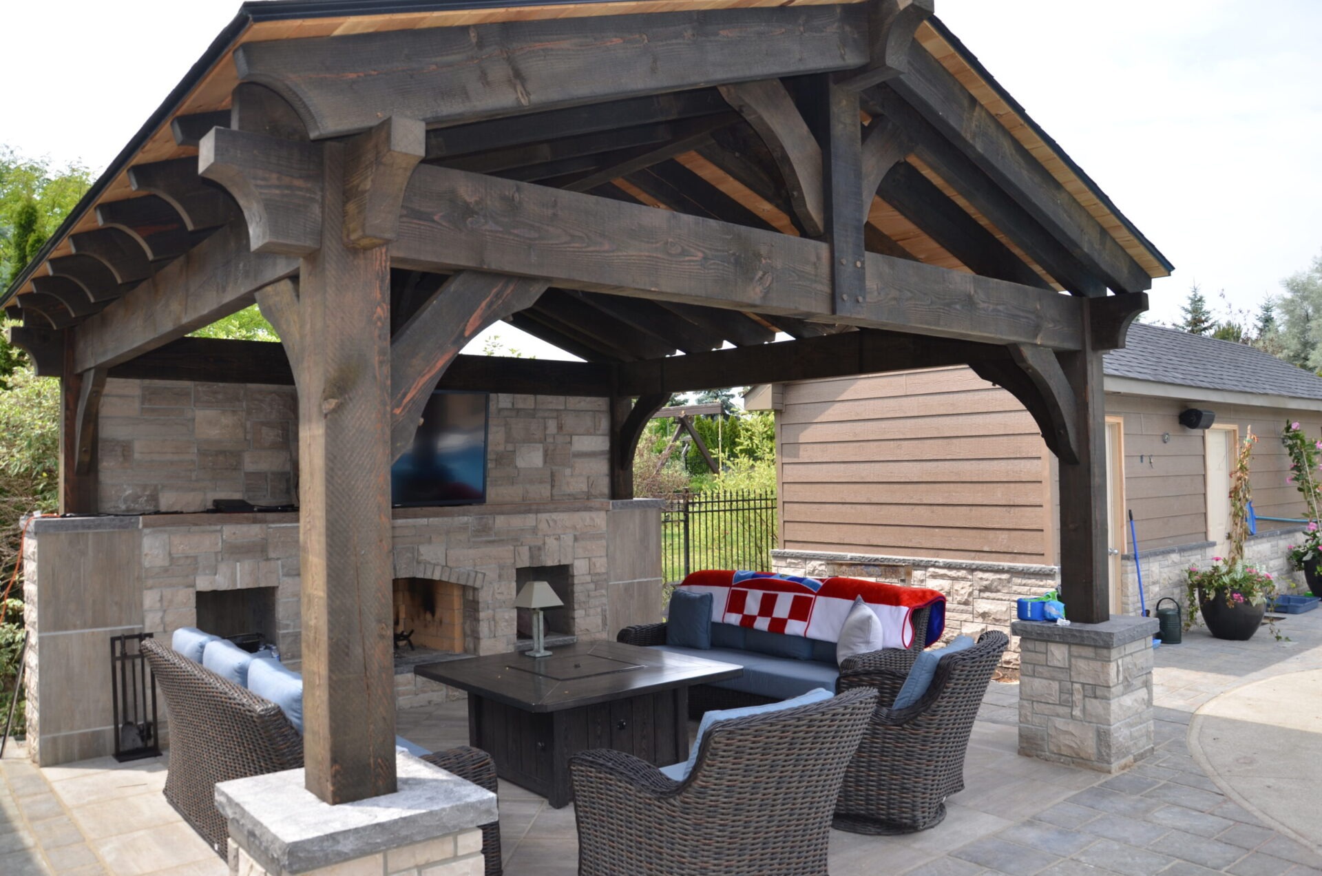 Outdoor patio with wooden pergola, stone fireplace, and wicker furniture. Vibrant red and white blanket on sofa enhances cozy atmosphere. Lush greenery surrounds area.