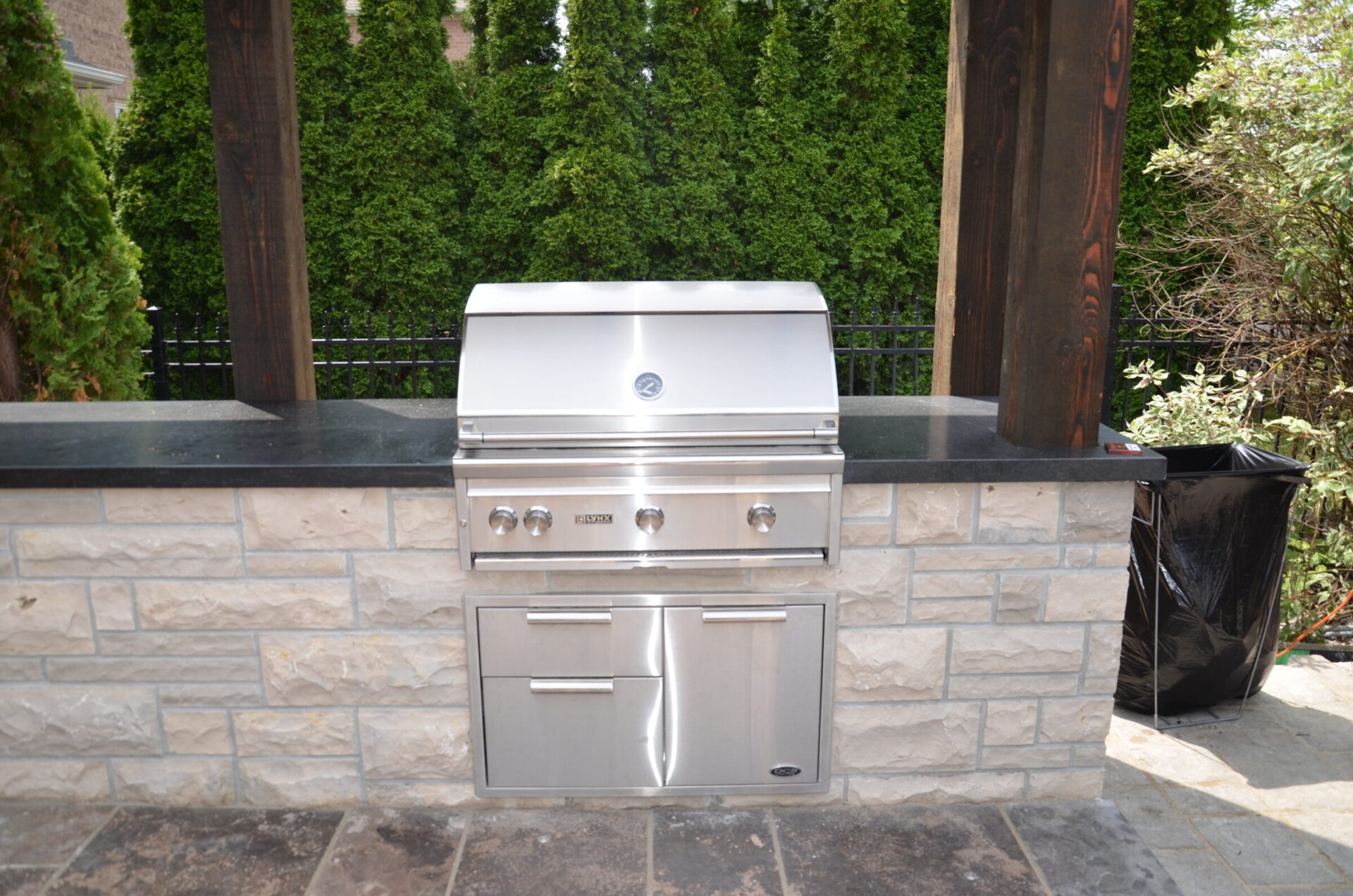 A stainless steel barbecue grill is set in a stone outdoor kitchen, surrounded by lush green hedges and natural stone tiles.