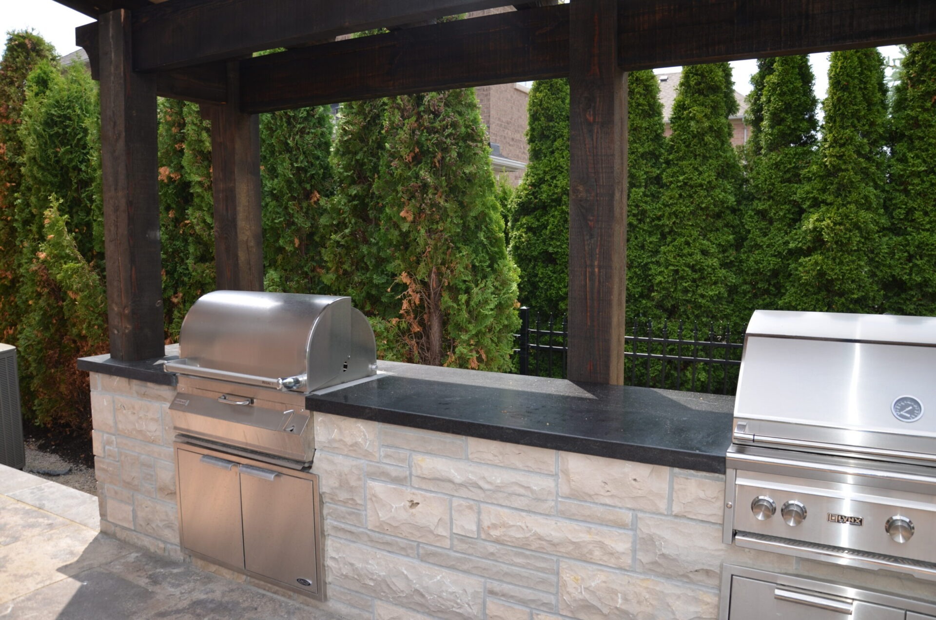 Outdoor kitchen with stone counter, stainless steel grills under a wooden pergola, surrounded by tall evergreen trees. Modern and sleek design.