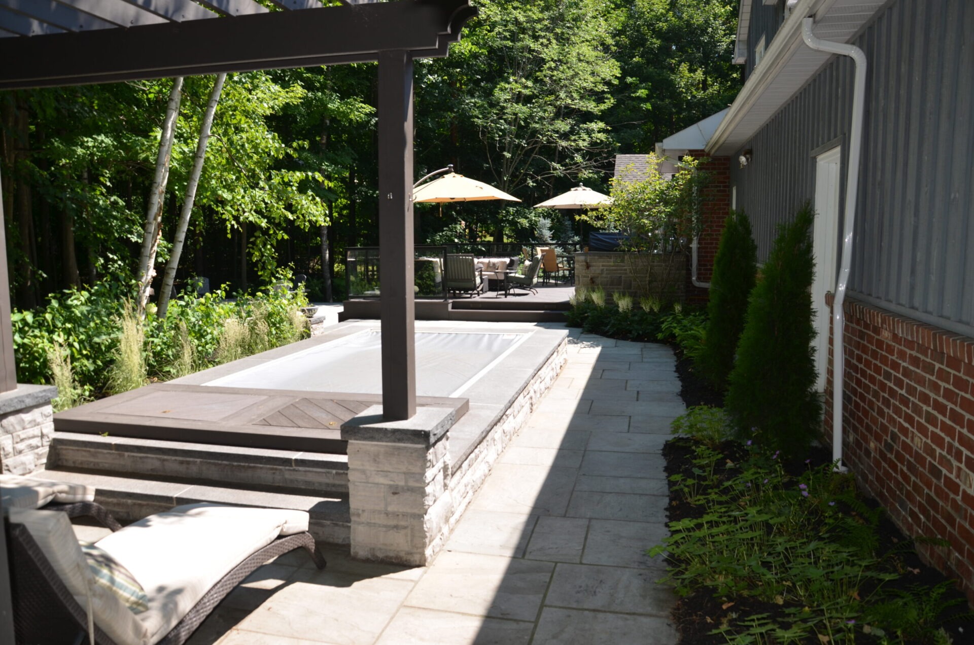 A modern backyard with a covered patio, swimming pool, and seating area under umbrellas, surrounded by lush greenery and trees.