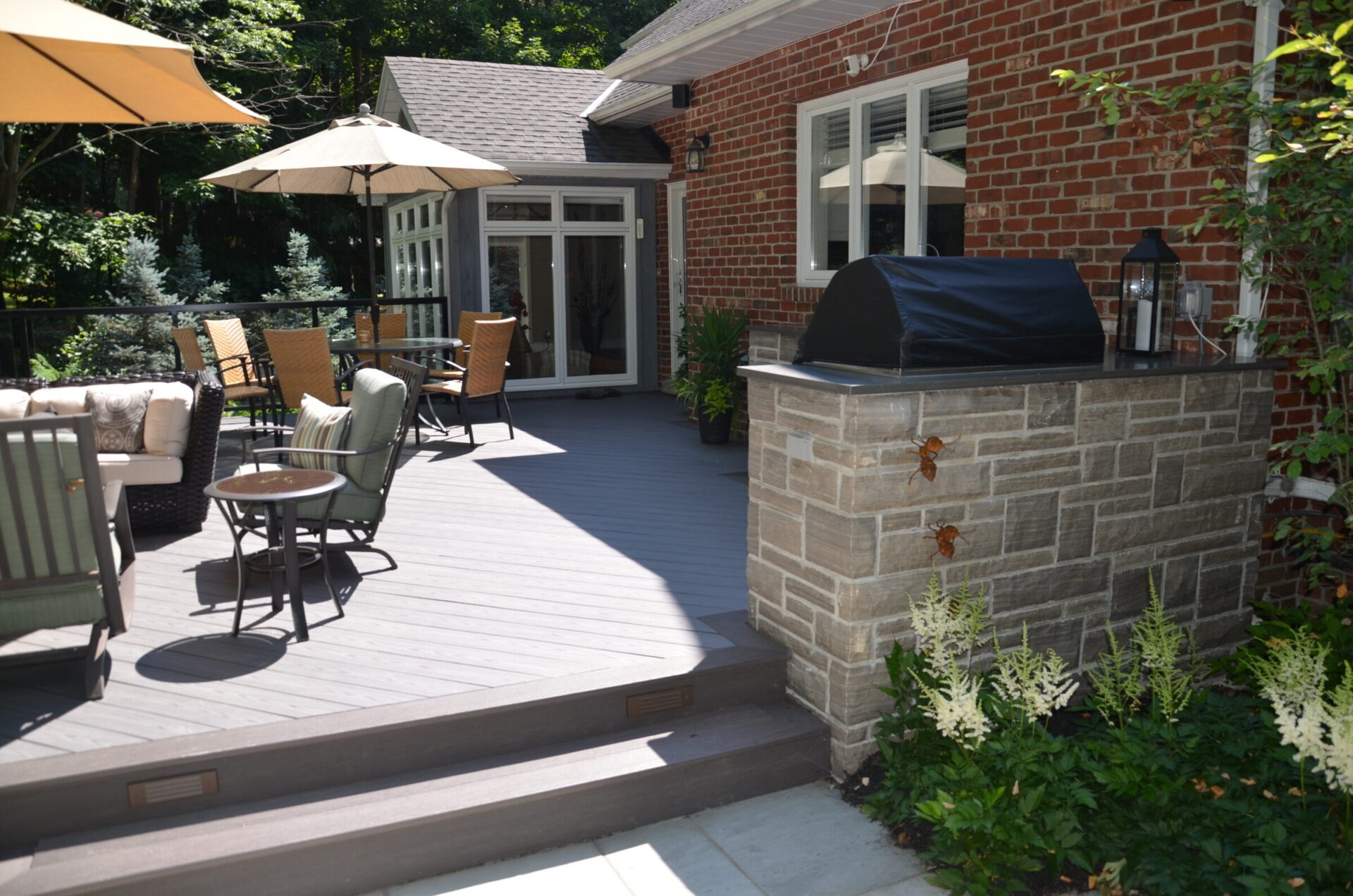 Spacious patio with outdoor furniture, umbrellas, and a grill. Surrounded by greenery, adjacent to a brick house with large windows.