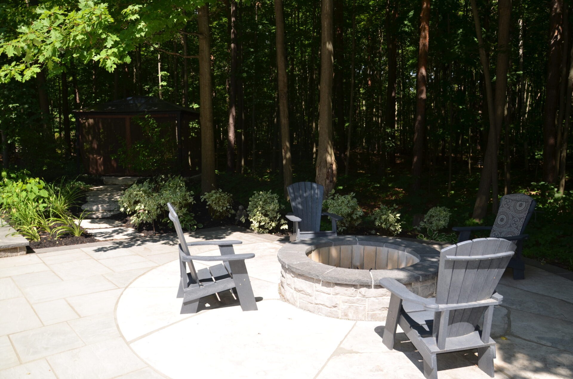 A serene patio scene with a stone fire pit, surrounded by Adirondack chairs and lush green forest in the background. Peaceful setting.