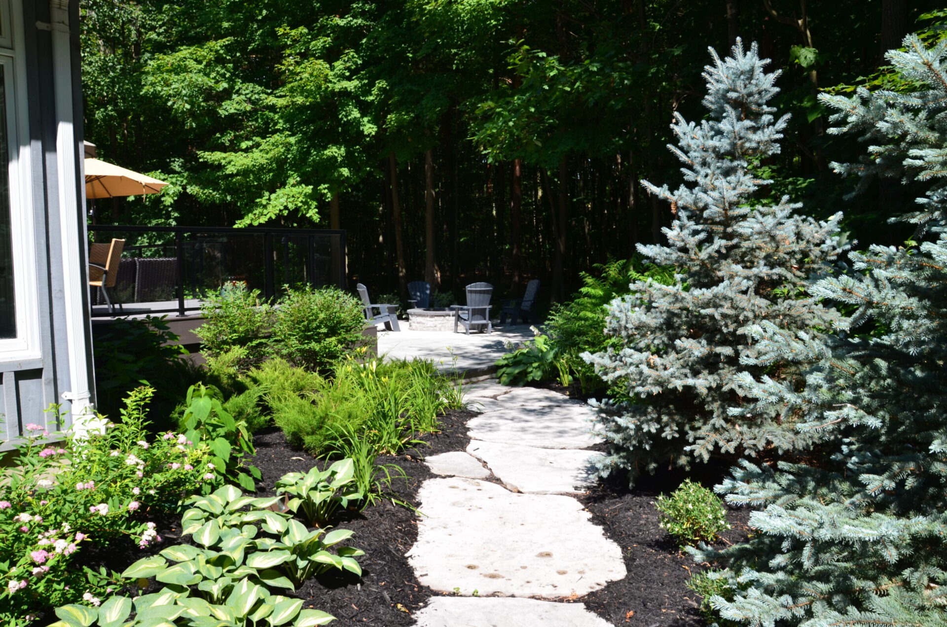 A lush garden path leads to a patio surrounded by trees and greenery, with chairs and a table visible under sunlight.