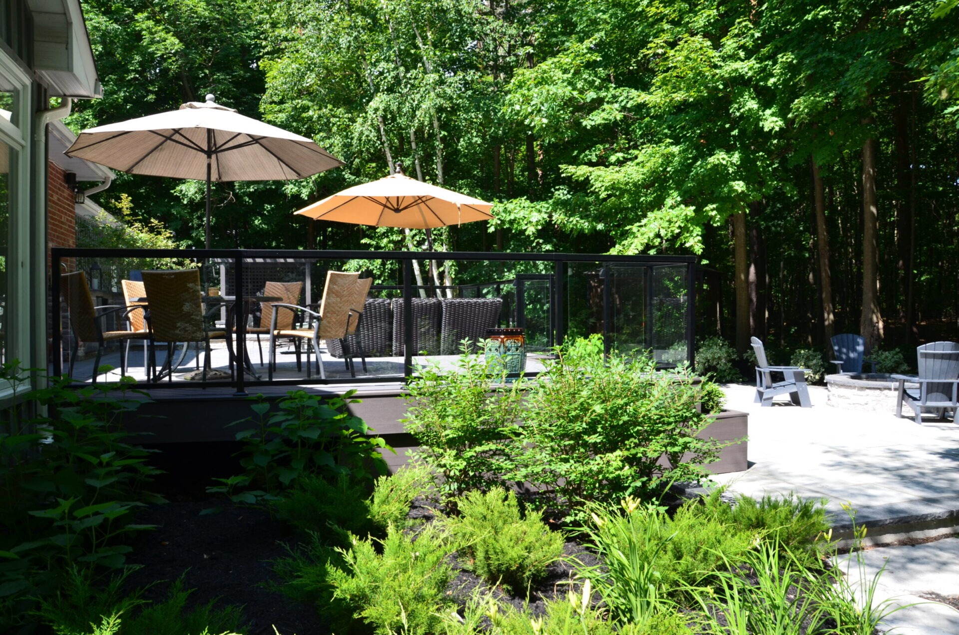 A sunlit patio with seating, umbrellas, and lush greenery. Surrounded by trees, it offers a serene outdoor space for relaxation and gatherings.