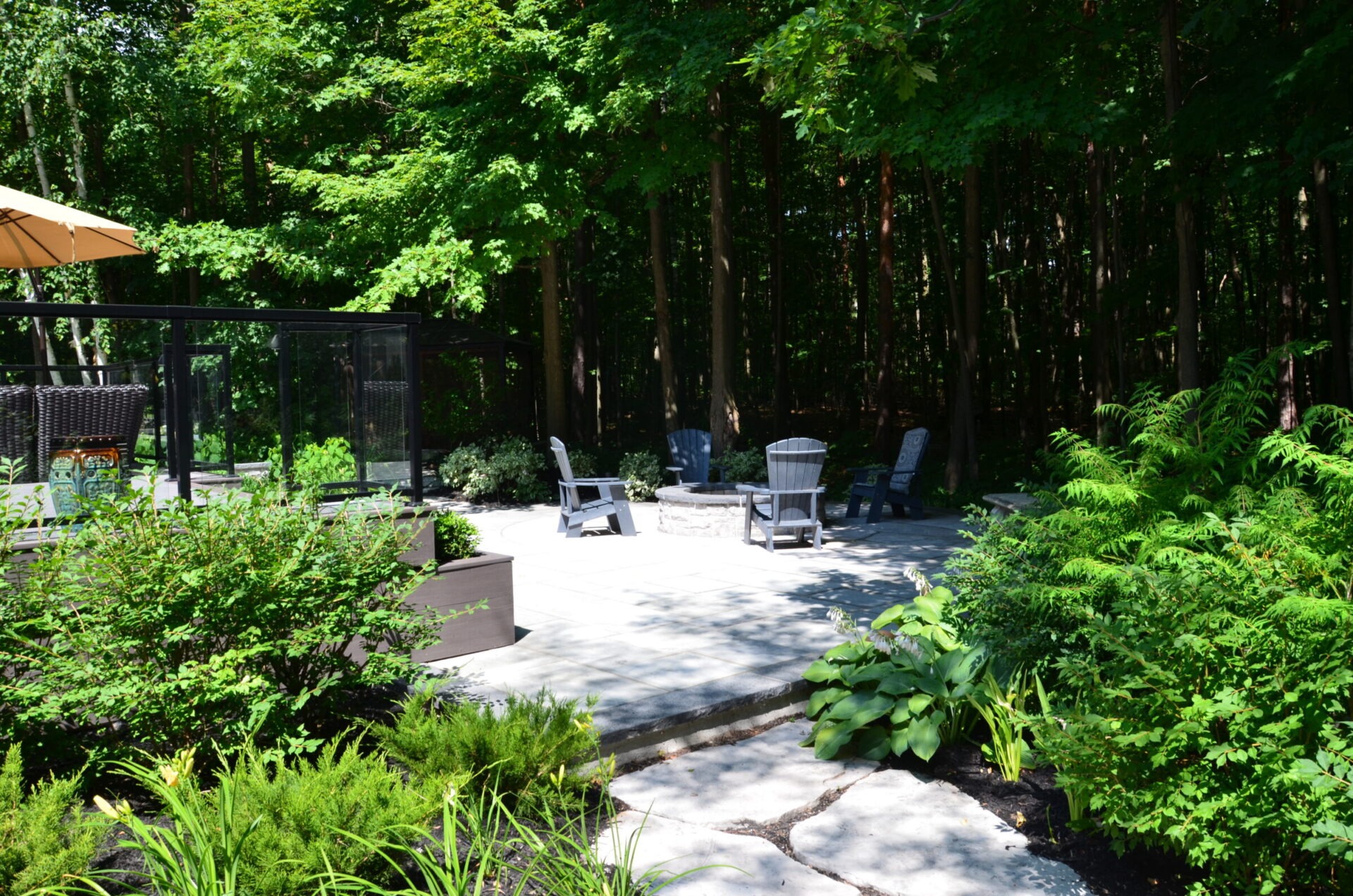 A serene patio set amidst lush greenery, featuring Adirondack chairs around a fire pit, bordered by dense forest and landscaped garden.