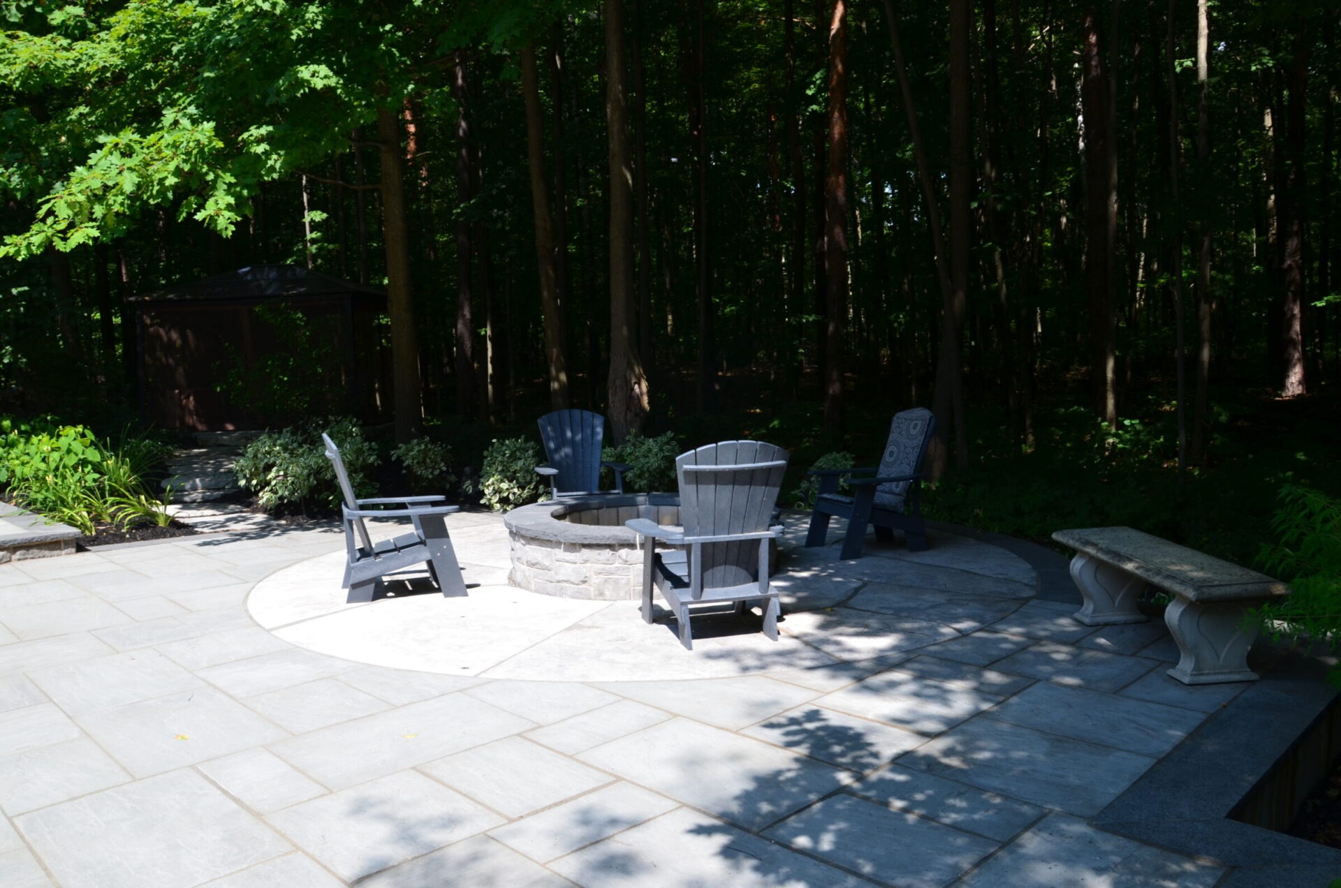 A stone patio with Adirondack chairs surrounds a fire pit, set against a backdrop of lush trees and greenery in a peaceful setting.