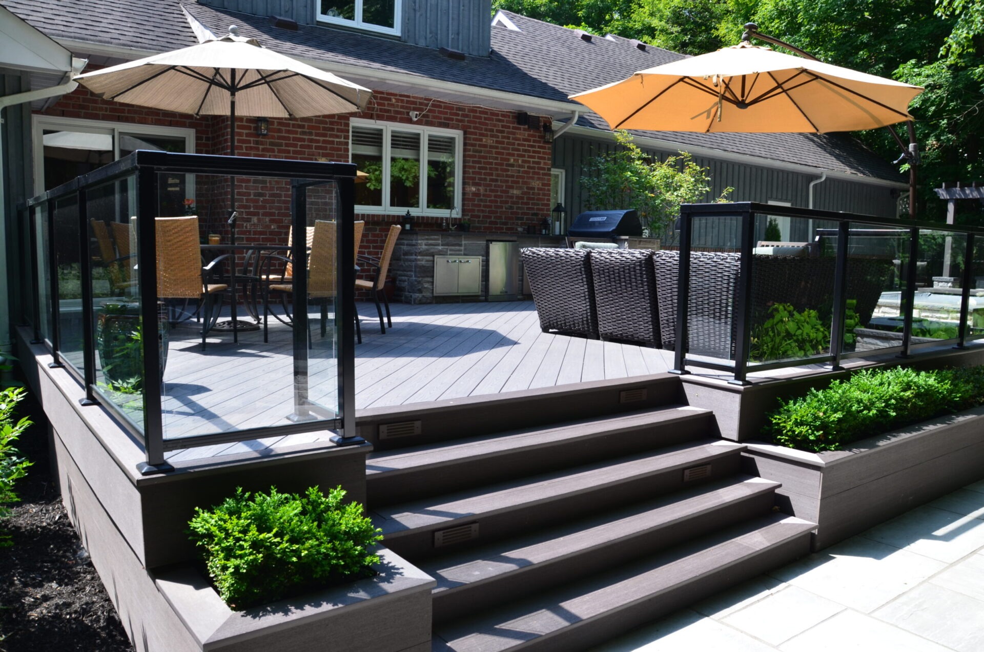 Modern patio with glass railing, wicker furniture, and umbrellas. Steps lead to lush garden. Brick house backdrop with large windows and greenery surrounding.