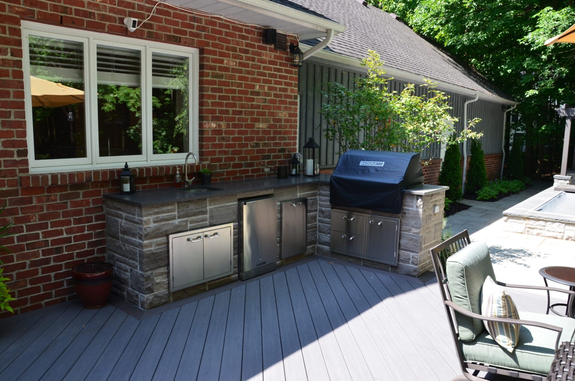 A cozy backyard patio features a stone outdoor kitchen with a grill, nestled beside a comfortable seating area and lush greenery.