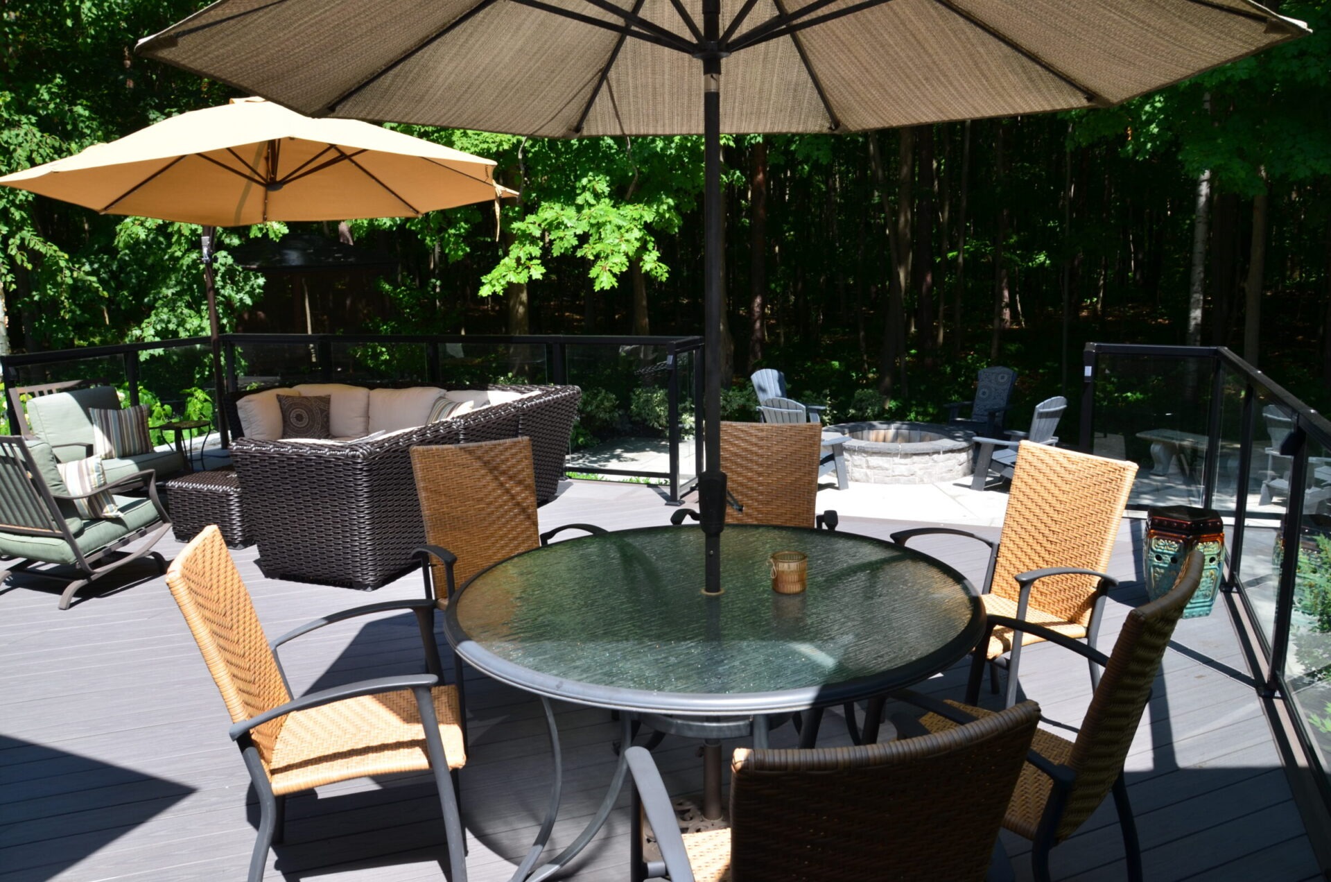 Outdoor patio with wicker furniture, glass table, and umbrellas. Surrounded by a forest backdrop, it includes a fire pit area with seating.