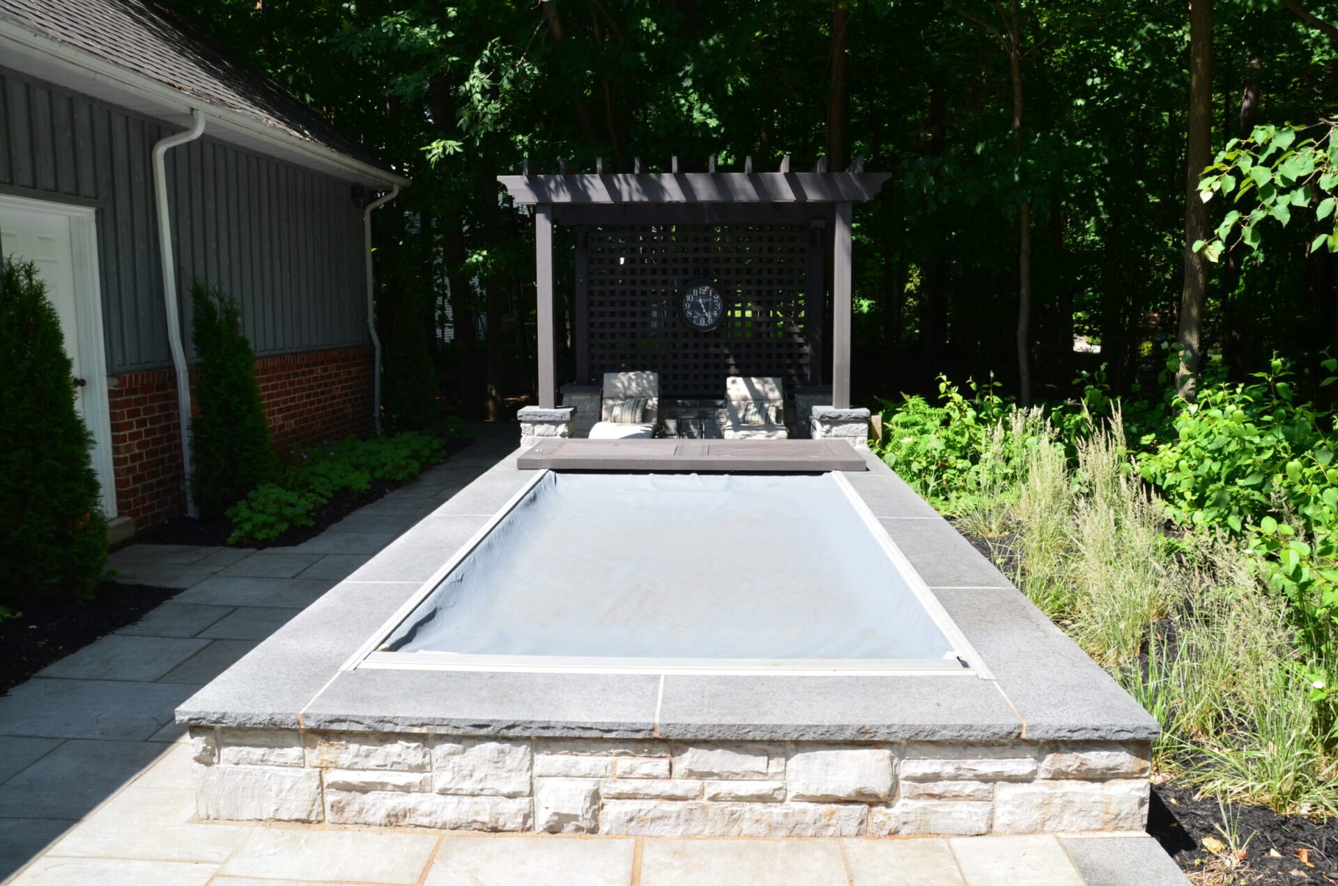 A small, covered pool surrounded by a stone patio, shaded by trees, with lounge chairs and a pergola in the background.