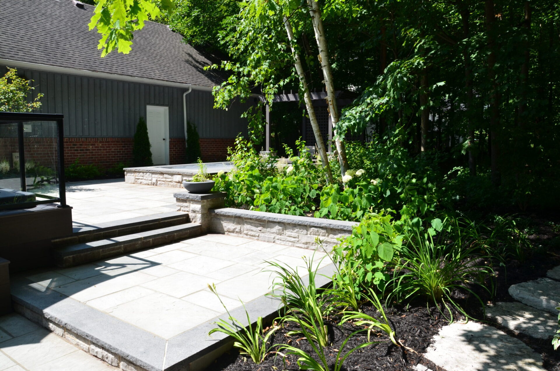 Modern outdoor patio with stone paving, raised planters, and lush greenery. Adjacent building features brick and gray siding. Tranquil and shaded setting.