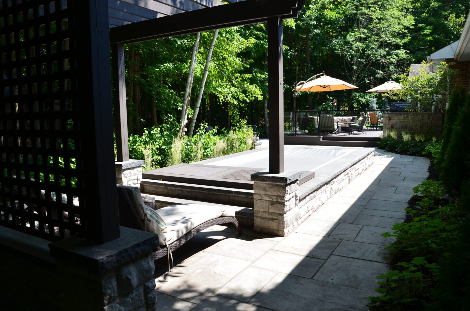 Lush garden area with a sunlit wooden pathway, lounge chairs, stone accents, and shaded seating under large umbrellas amidst green foliage.