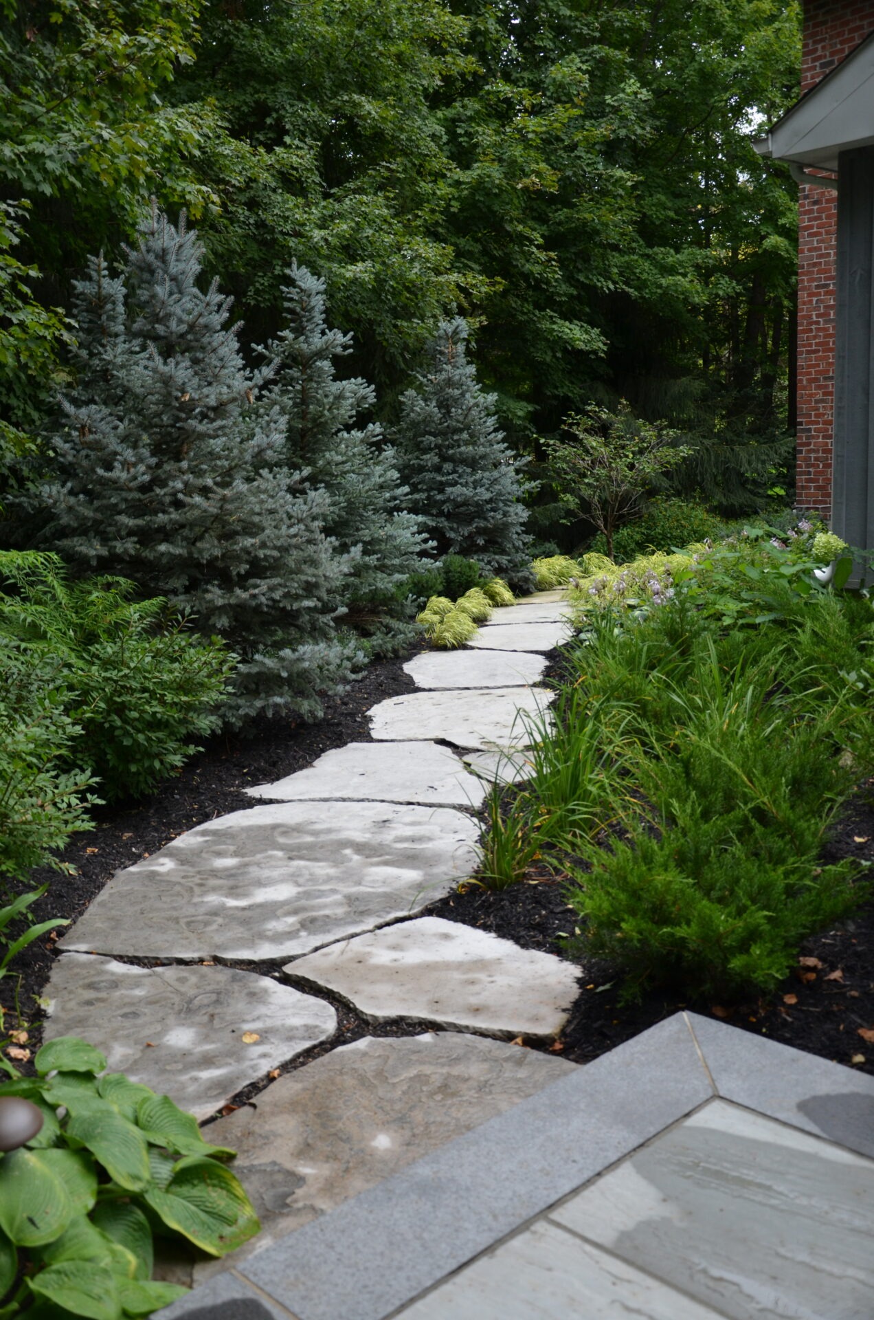 A stone path winds through a lush, green garden with various plants and trees, next to a brick building wall.