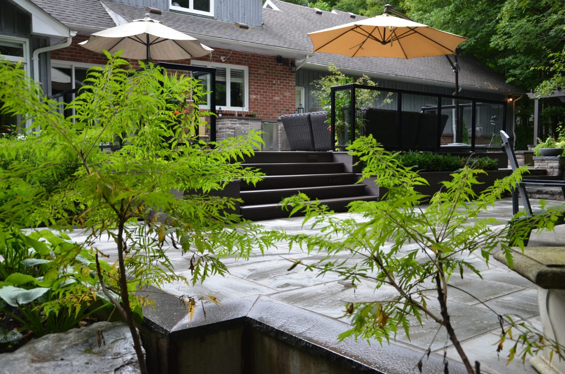 A cozy backyard with patio furniture, umbrellas, lush greenery, and a brick house in the background, creating a serene outdoor atmosphere.