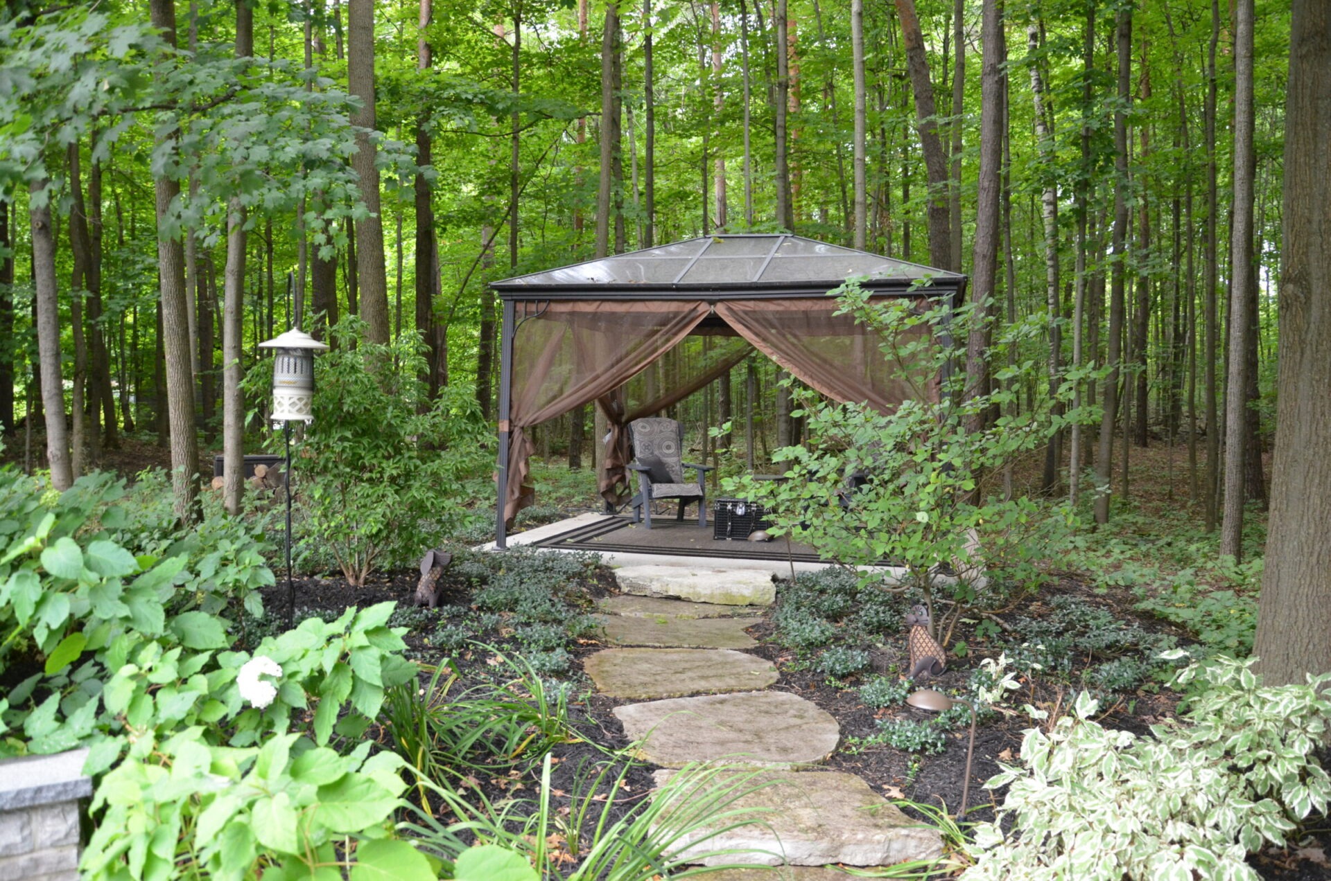 A tranquil garden scene with a gazebo surrounded by lush greenery, a stone path leading to a cozy outdoor seating area.