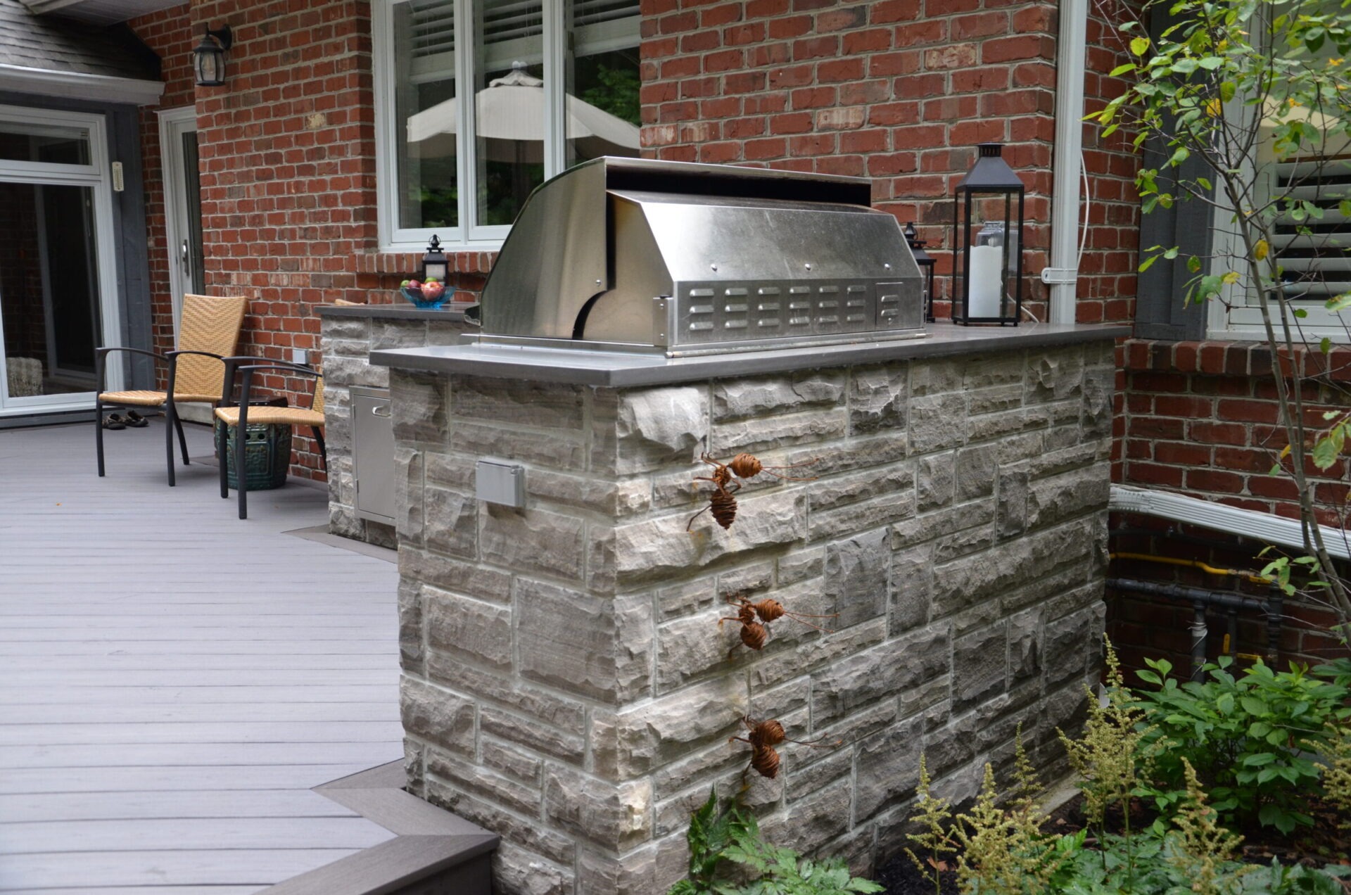 A cozy backyard patio features a stone barbecue grill, wicker chairs, and potted plants against a red brick house backdrop.