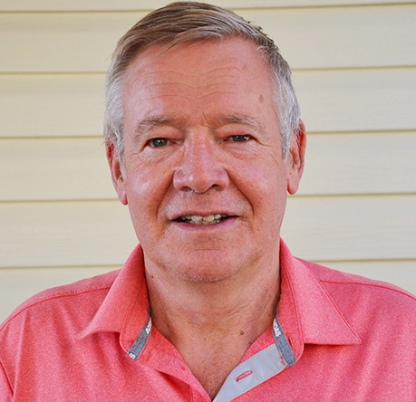 A person with gray hair wearing a pink shirt stands in front of a light yellow wall. The background is plain and simple.