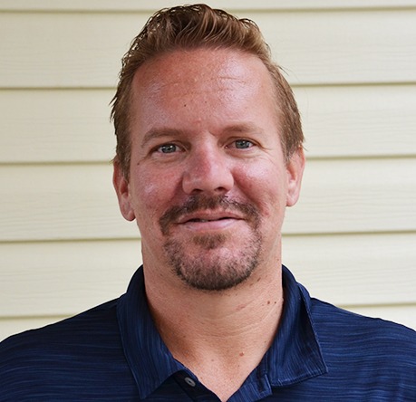 A person with short hair and a goatee smiles slightly, standing against a light-colored, horizontal panel wall. Casual navy-colored shirt worn.