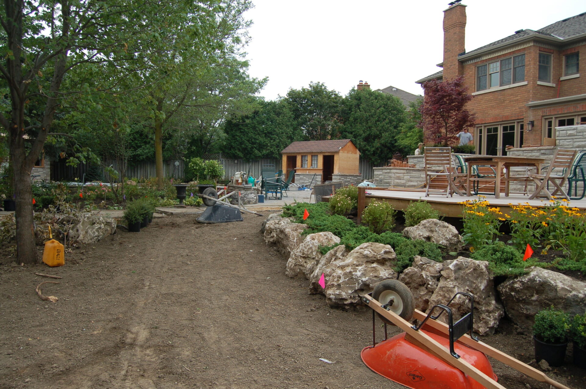 Backyard under construction with rocks, wheelbarrow, and potted plants. Wooden deck with furniture, trees, and shrubs in the background. No people visible.