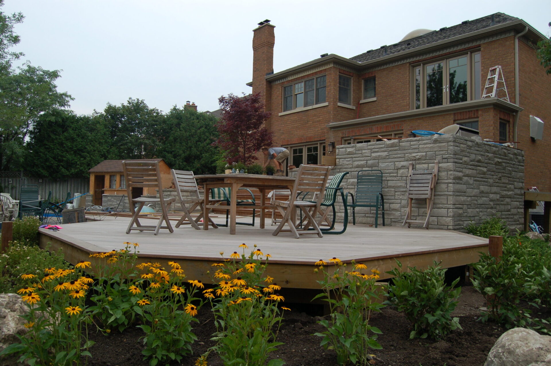 A person stands on a wooden deck in a backyard with chairs, a brick house, and surrounding garden. Overcast sky in the background.