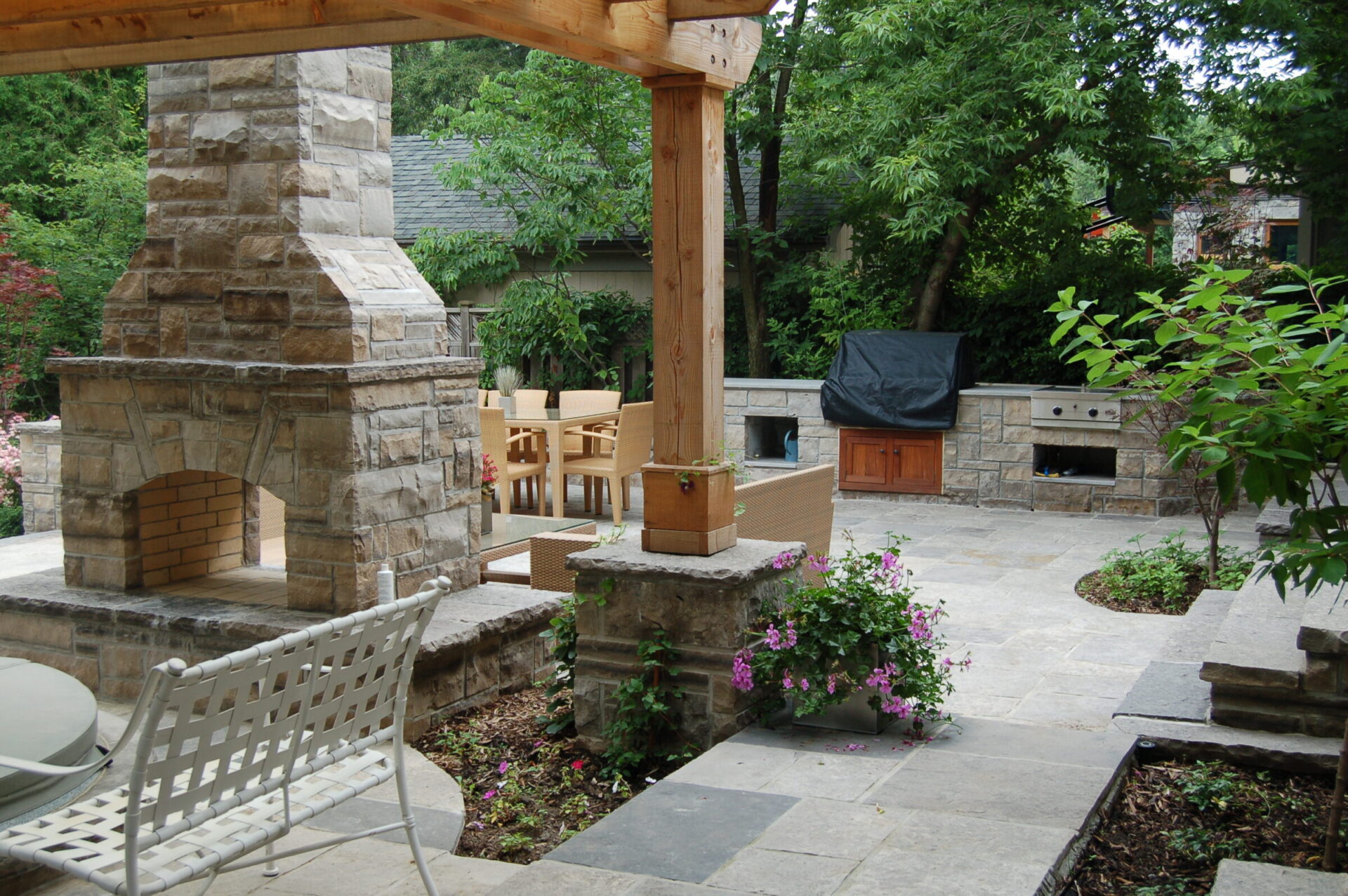 Elegant backyard features stone fireplace, dining area, and green foliage, creating a cozy outdoor living space in a serene garden setting.