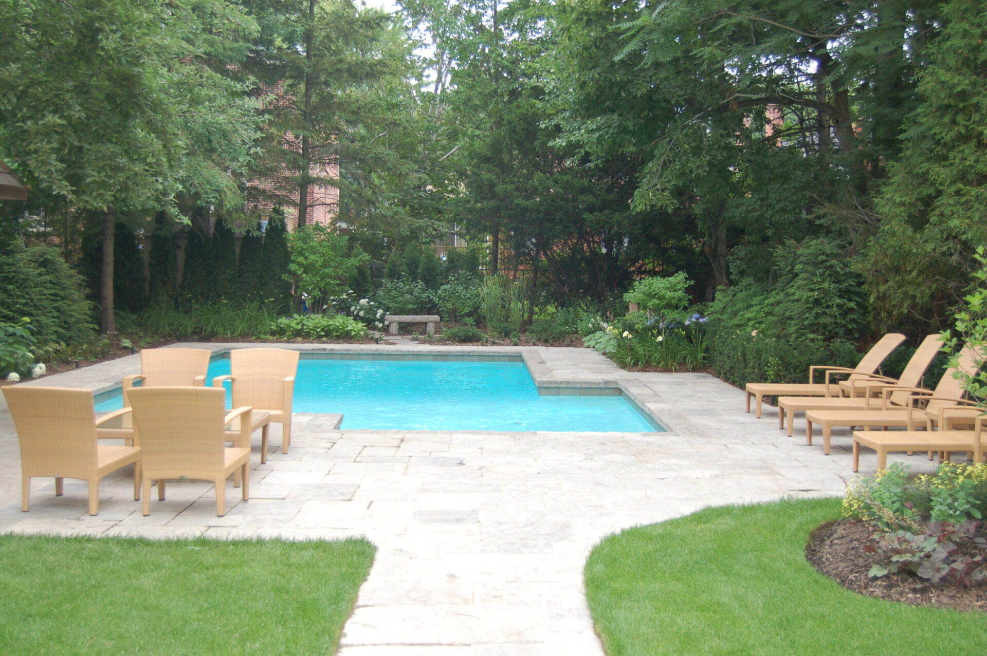 A serene backyard with a rectangular pool, surrounded by lush greenery. Wooden lounge chairs and a seating area provide relaxation spots.