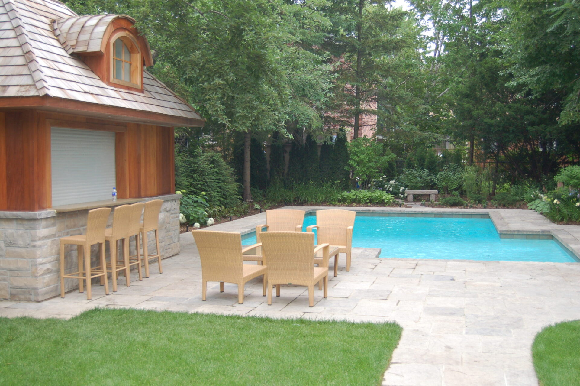 A serene backyard featuring a pool, wooden patio chairs, and a small bar structure, surrounded by lush greenery and trees.