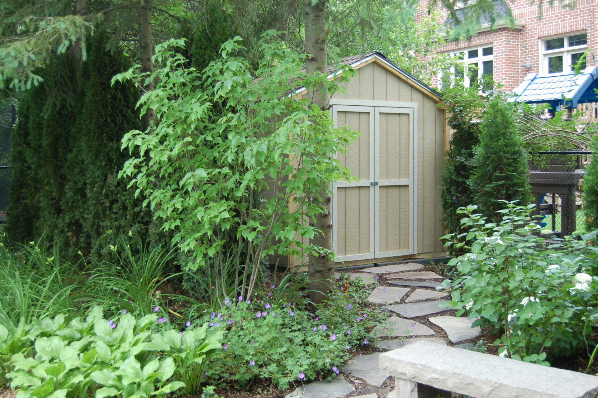 A small garden shed is surrounded by bushes, stone path, and trees, with a brick building visible in the background.