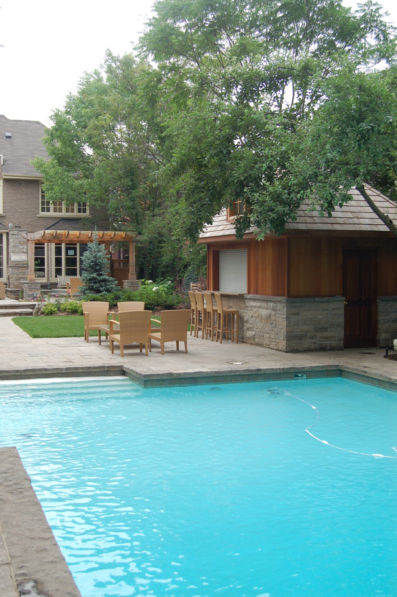 A serene backyard scene with a pool, wooden patio furniture, and a stone outbuilding surrounded by lush trees and greenery.