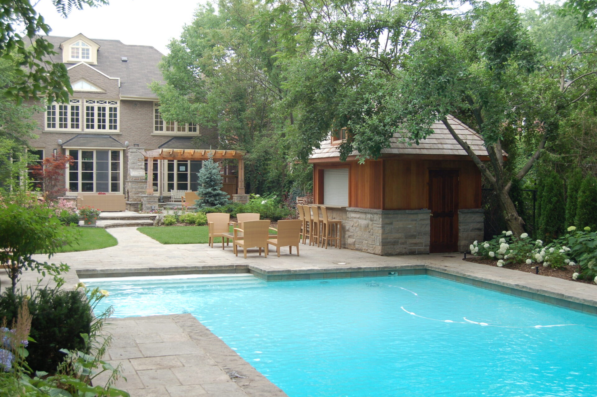 A backyard features a swimming pool, patio furniture, and a small poolside building surrounded by trees and greenery, attached to a large house.