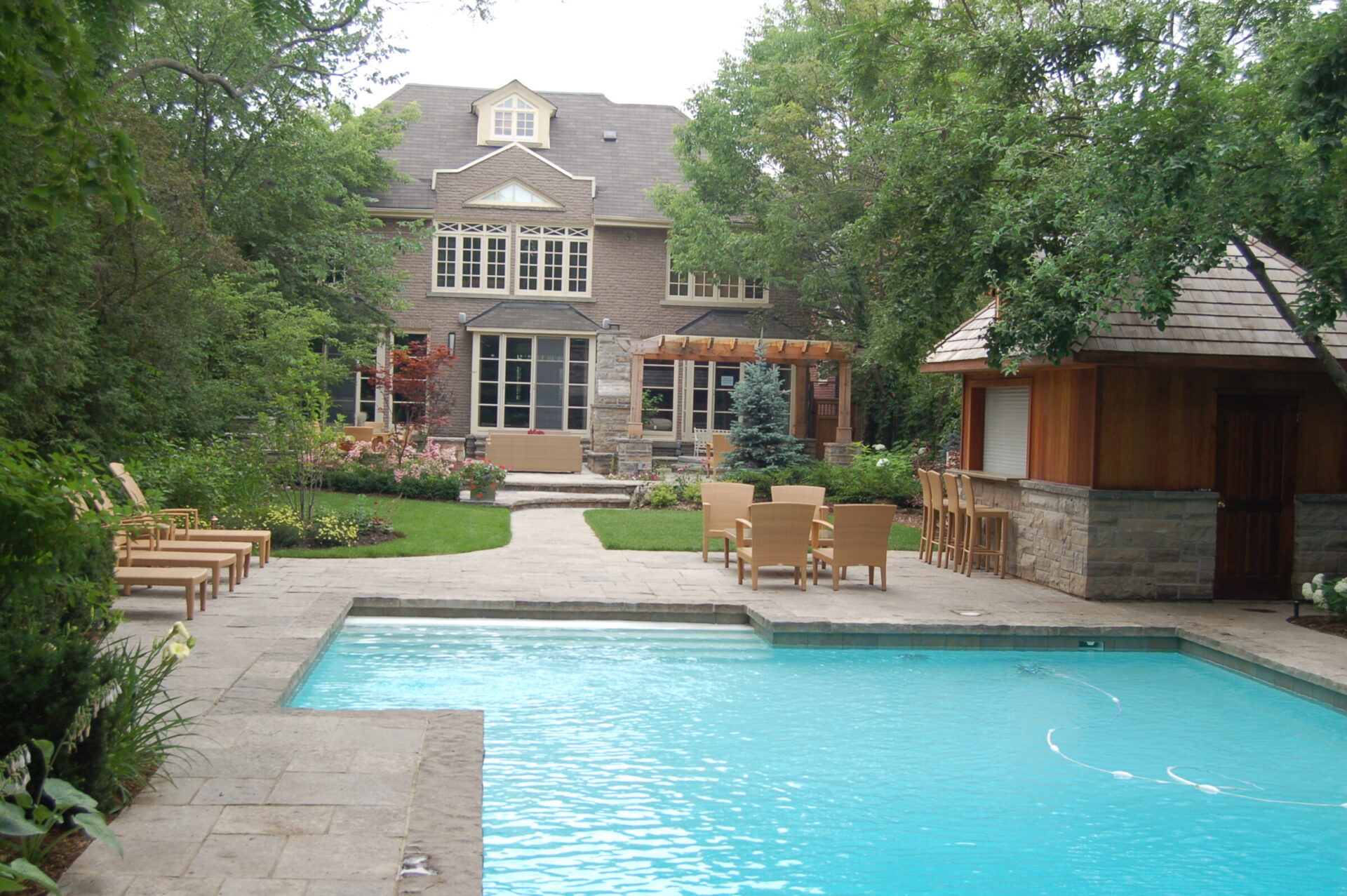 A spacious backyard features a swimming pool, patio chairs, and a wooden pool house, surrounded by lush greenery and a large brick house.