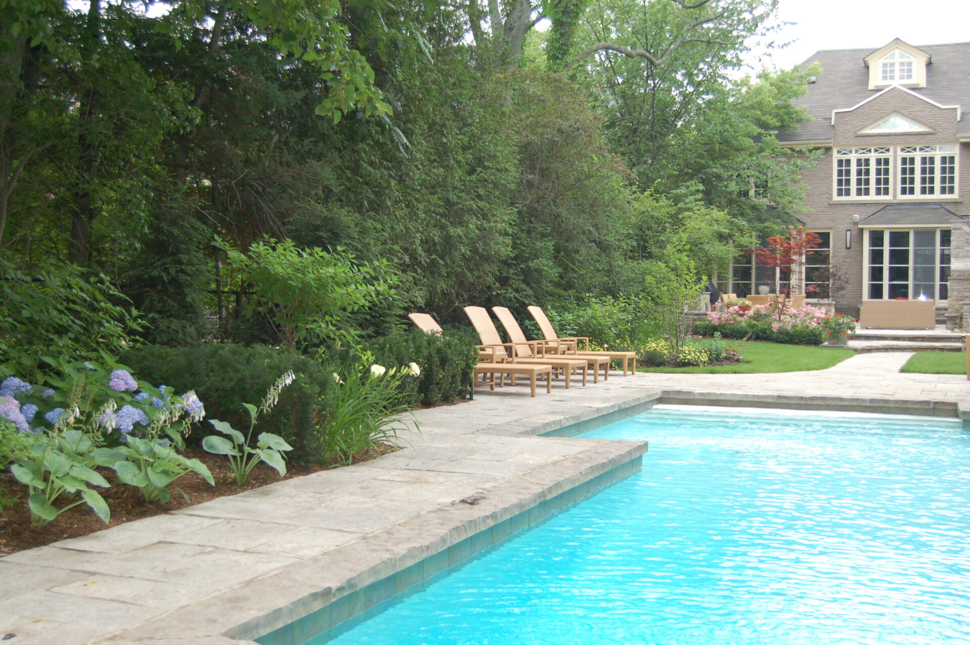 A serene backyard features a clear swimming pool, surrounded by lush greenery and flowers, with lounge chairs and a large house in the background.