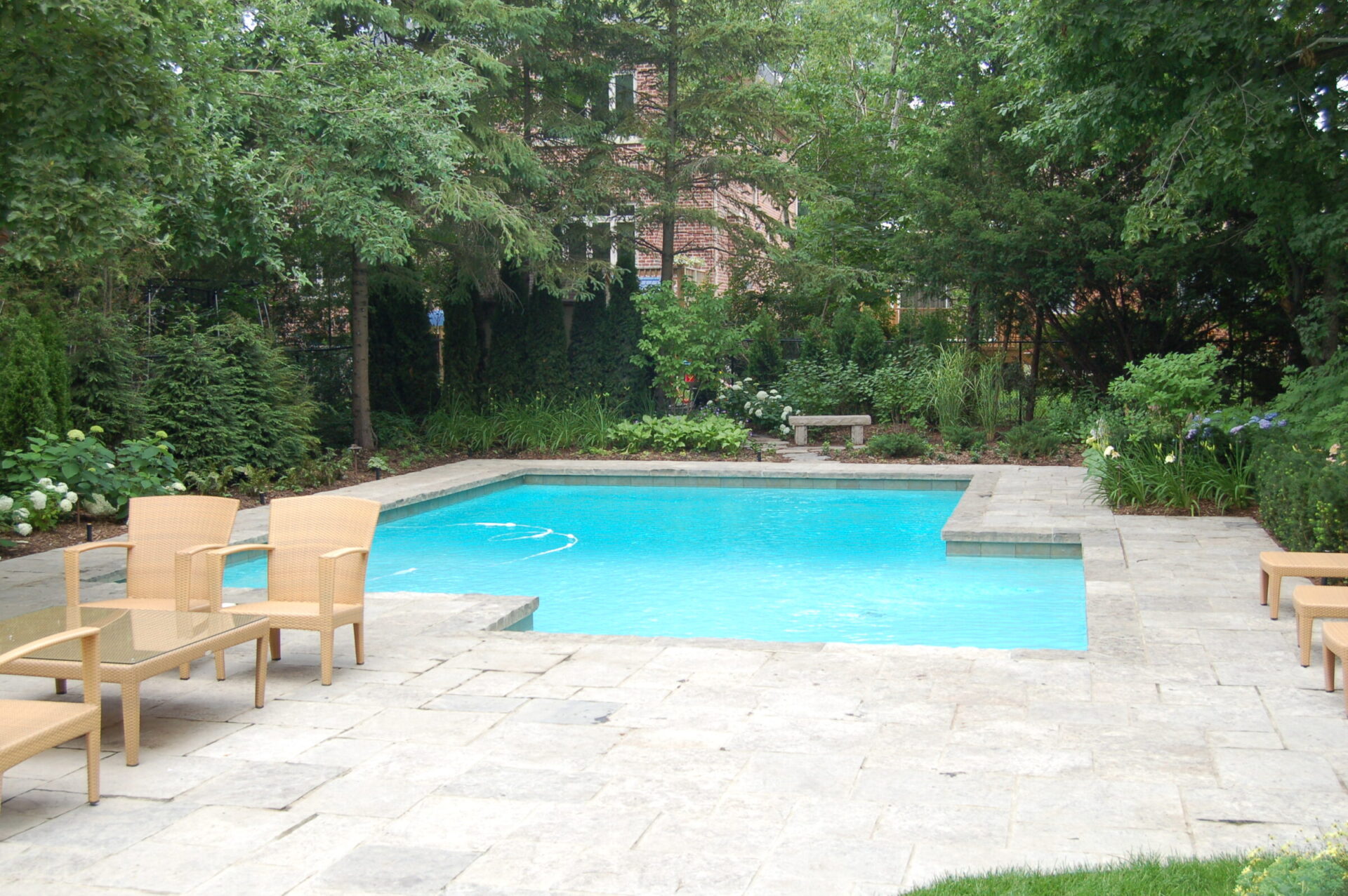 A serene backyard features a clear blue swimming pool, surrounded by lush greenery and outdoor furniture on a stone patio. No people visible.