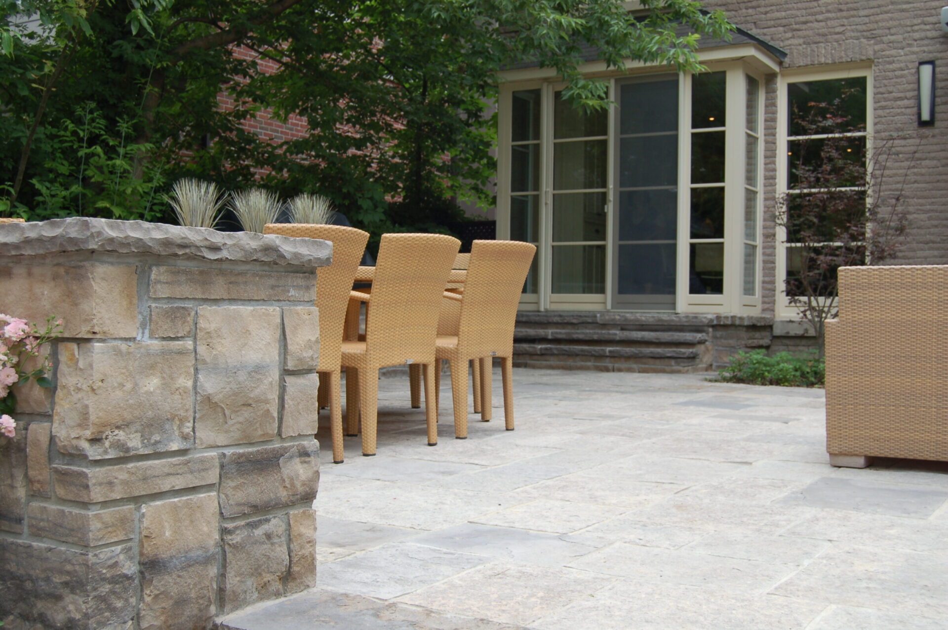 Outdoor patio with wicker chairs and a stone table, surrounded by lush greenery. Glass doors lead into a modern building.