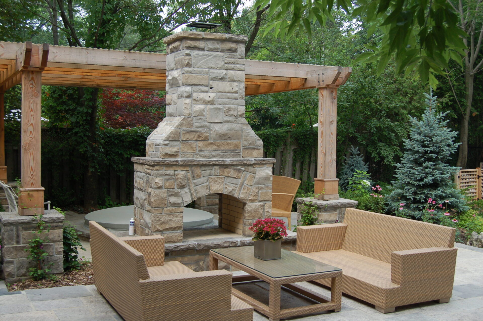 A cozy outdoor patio features a stone fireplace, wicker furniture, and a wooden pergola, surrounded by lush greenery and blooming flowers.