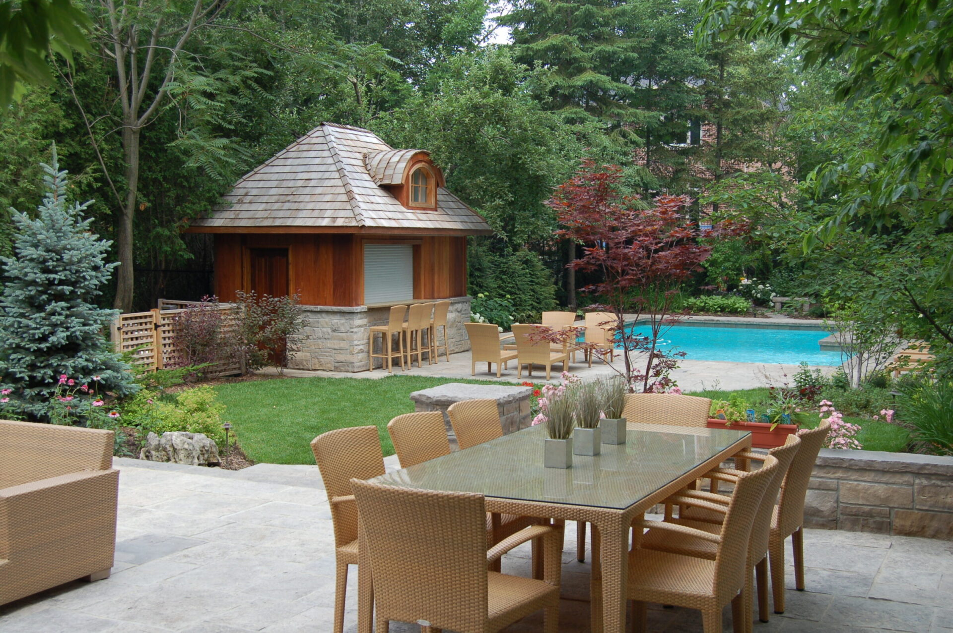 A tranquil backyard features a small wooden house, patio furniture, lush greenery, and a swimming pool, creating a peaceful outdoor setting.