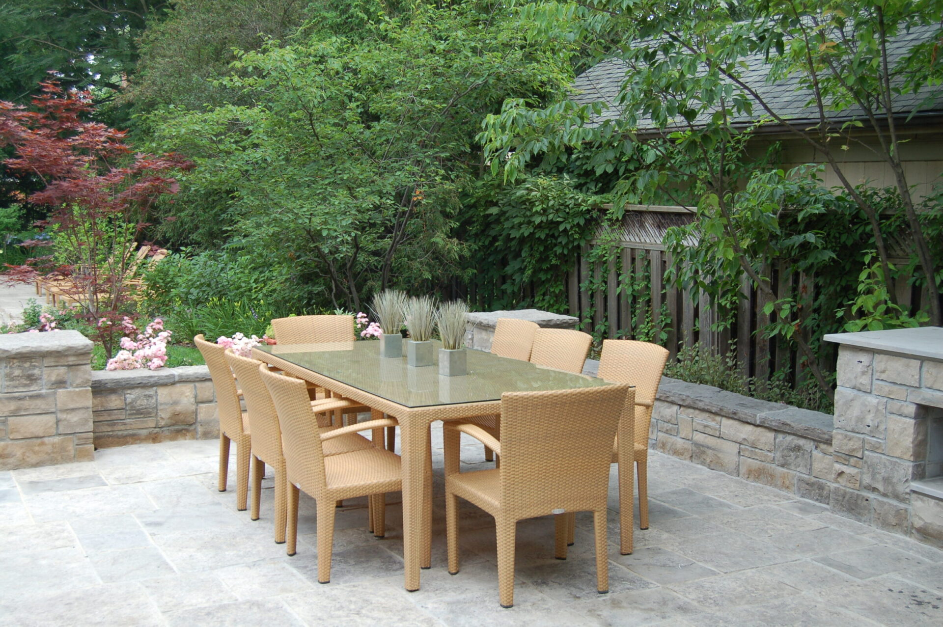 Outdoor patio with a glass table, wicker chairs, surrounded by lush greenery, stone flooring, and a wooden fence, creating a tranquil garden setting.